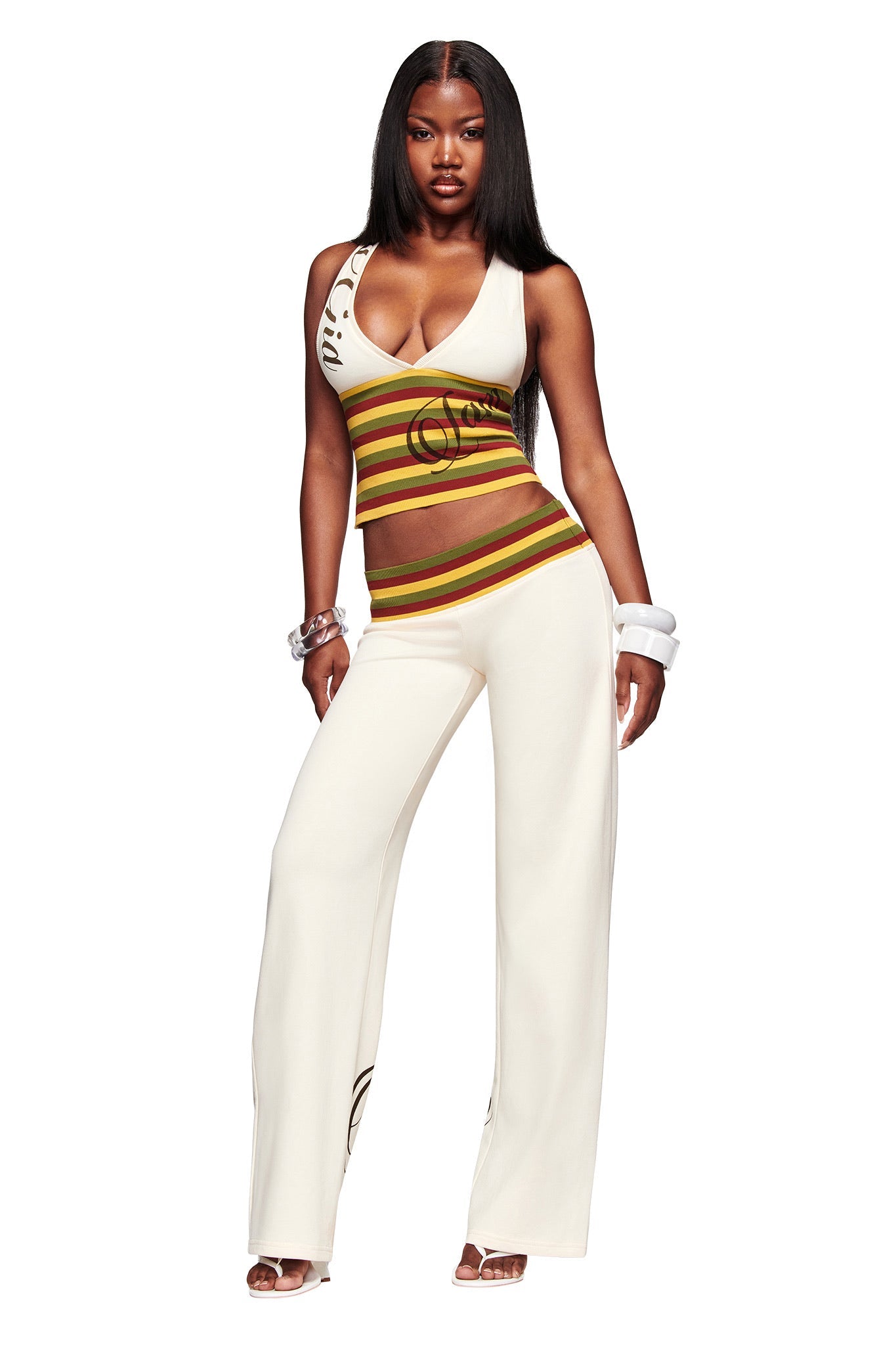 A woman stands confidently in the I.AM.GIA CANDI TOP - CREAM, a V-neck racerback with tri-coloured striped ribbing, paired with matching wide-leg white pants, chunky bracelets, and open-toe heels.