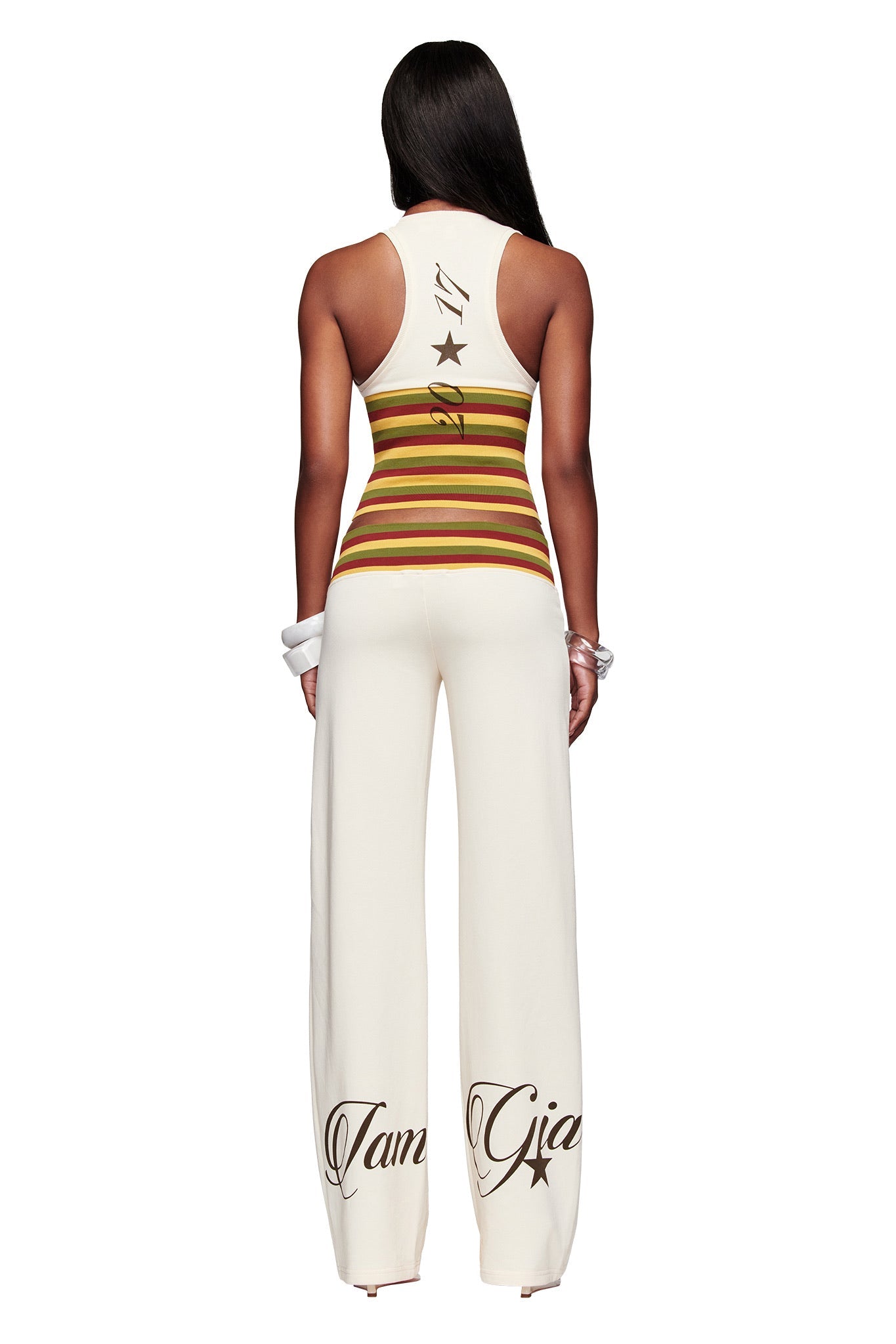 A woman with long dark hair faces away, wearing the I.AM.GIA CANDI TOP in cream—a sleeveless piece with tri-coloured striped ribbing at the waist and black lettering with stars on the back.