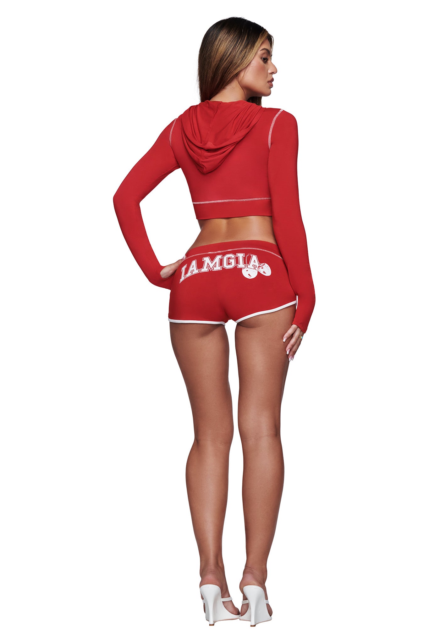 A woman stands with her back to the camera, wearing a red crop hoodie and I.AM.GIA's BLARE DOLPHIN SHORT in red, styled in a mini dolphin cut and paired with white high-heeled shoes.