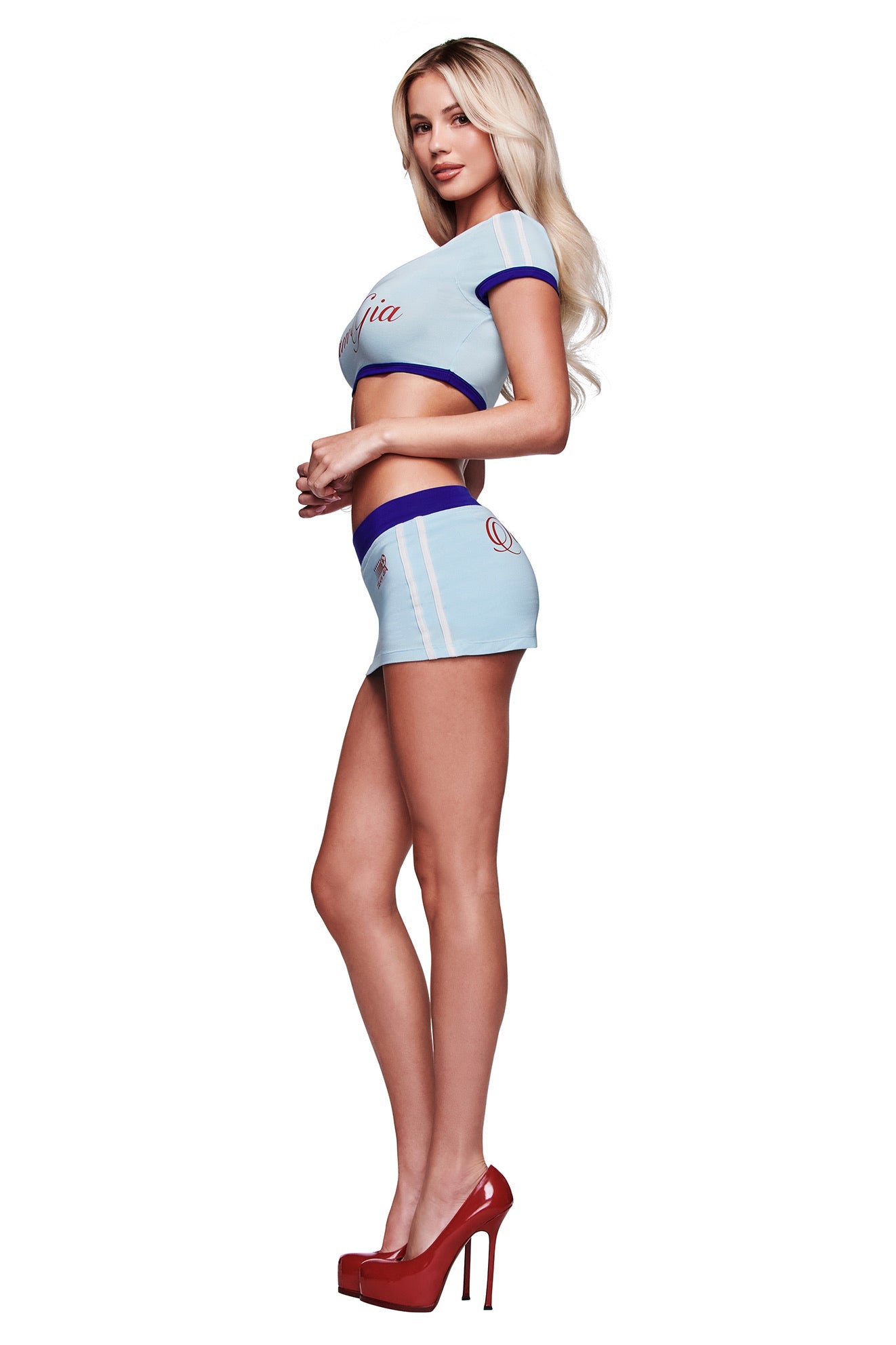 A woman with long blonde hair wears the I.AM.GIA BLARE SPORT CROP TEE AND HEADBAND in baby blue, paired with a matching mini skirt and red high heels, standing in profile against a white background.