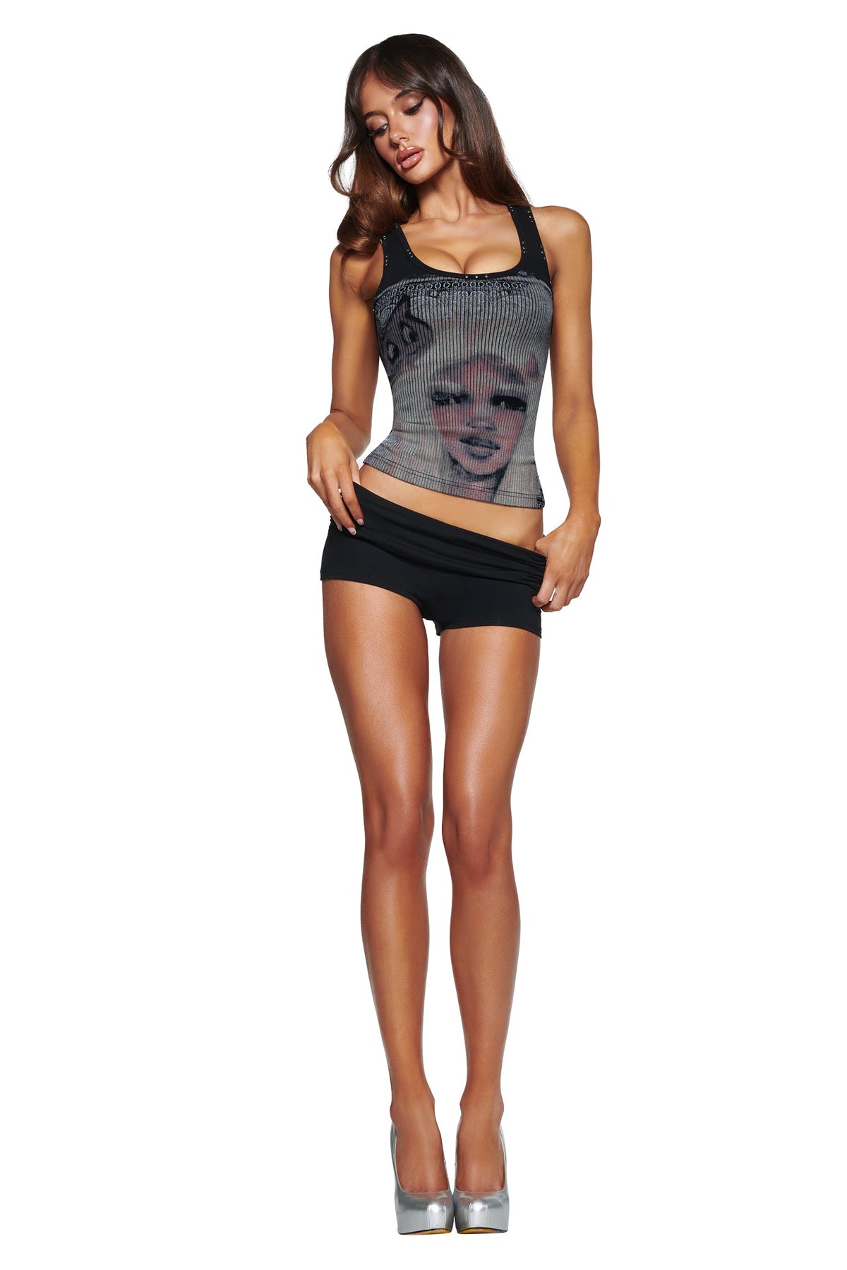 A woman with long brown hair wears the I.AM.GIA T-REX INA TANK - BLACK and black shorts, standing on silver high heels against a white background.