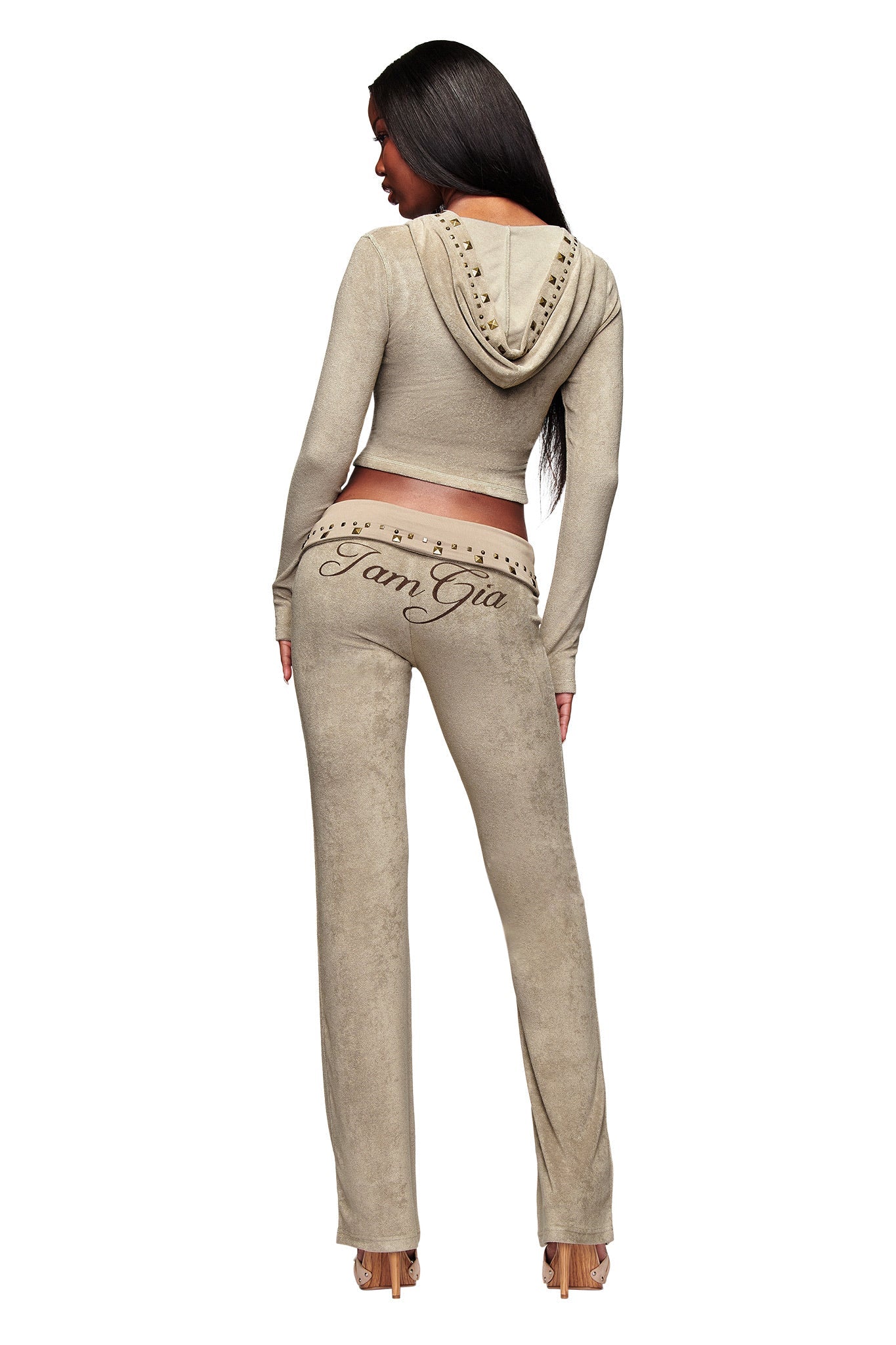 A woman faces away, wearing a tan hooded crop top and I.AM.GIA’s BLARE STUDDED TERRY PANT in Stone, featuring "I Am Gia" branding on the waistband and back. The studded terry cloth pants are styled with heels against a white backdrop.