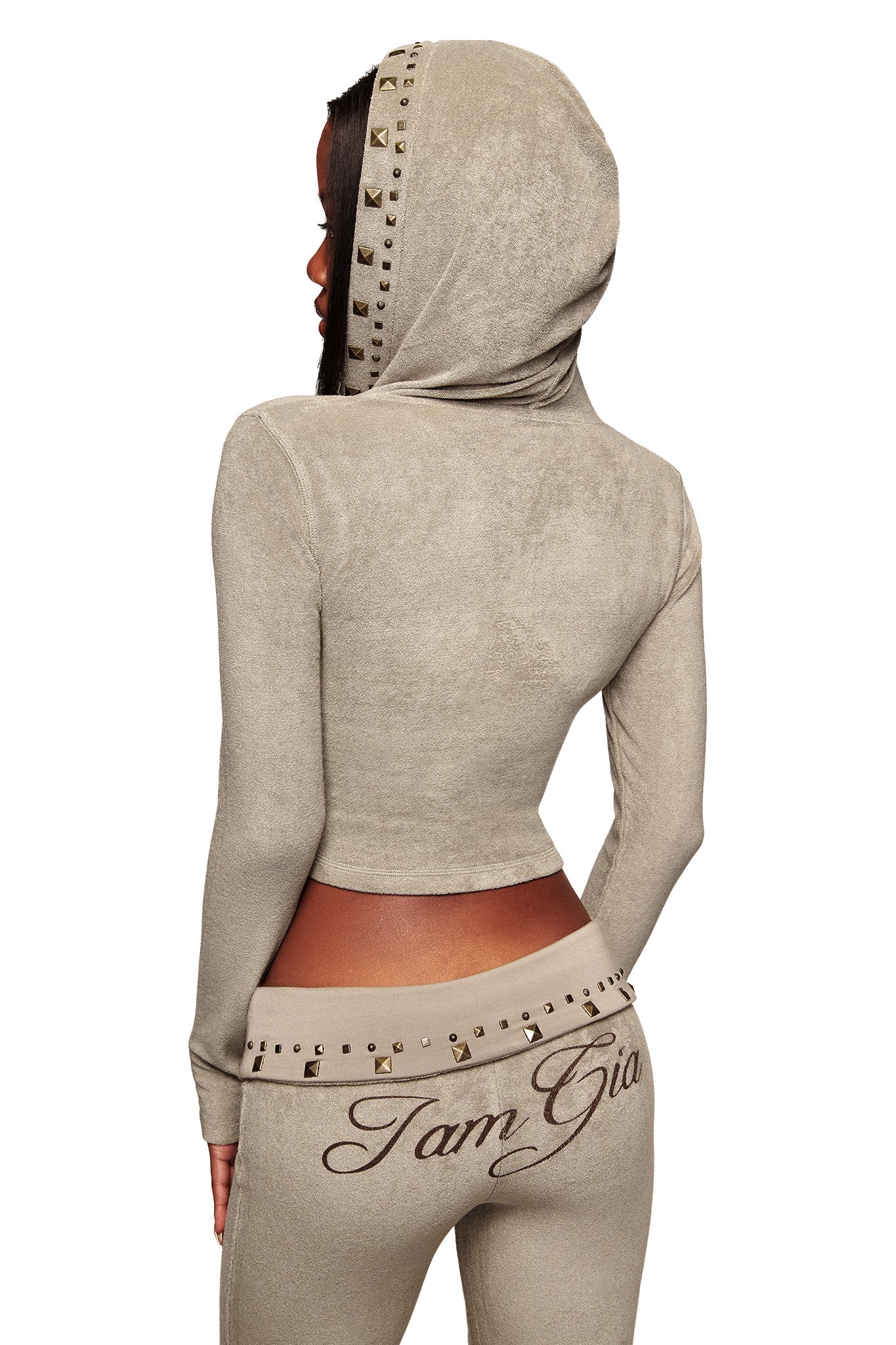 A woman faces away wearing the I.AM.GIA BLARE STUDDED TERRY HOODIE in stone, paired with matching pants. The outfit features studded accents and "Iam Gia" printed across the back of the pants.