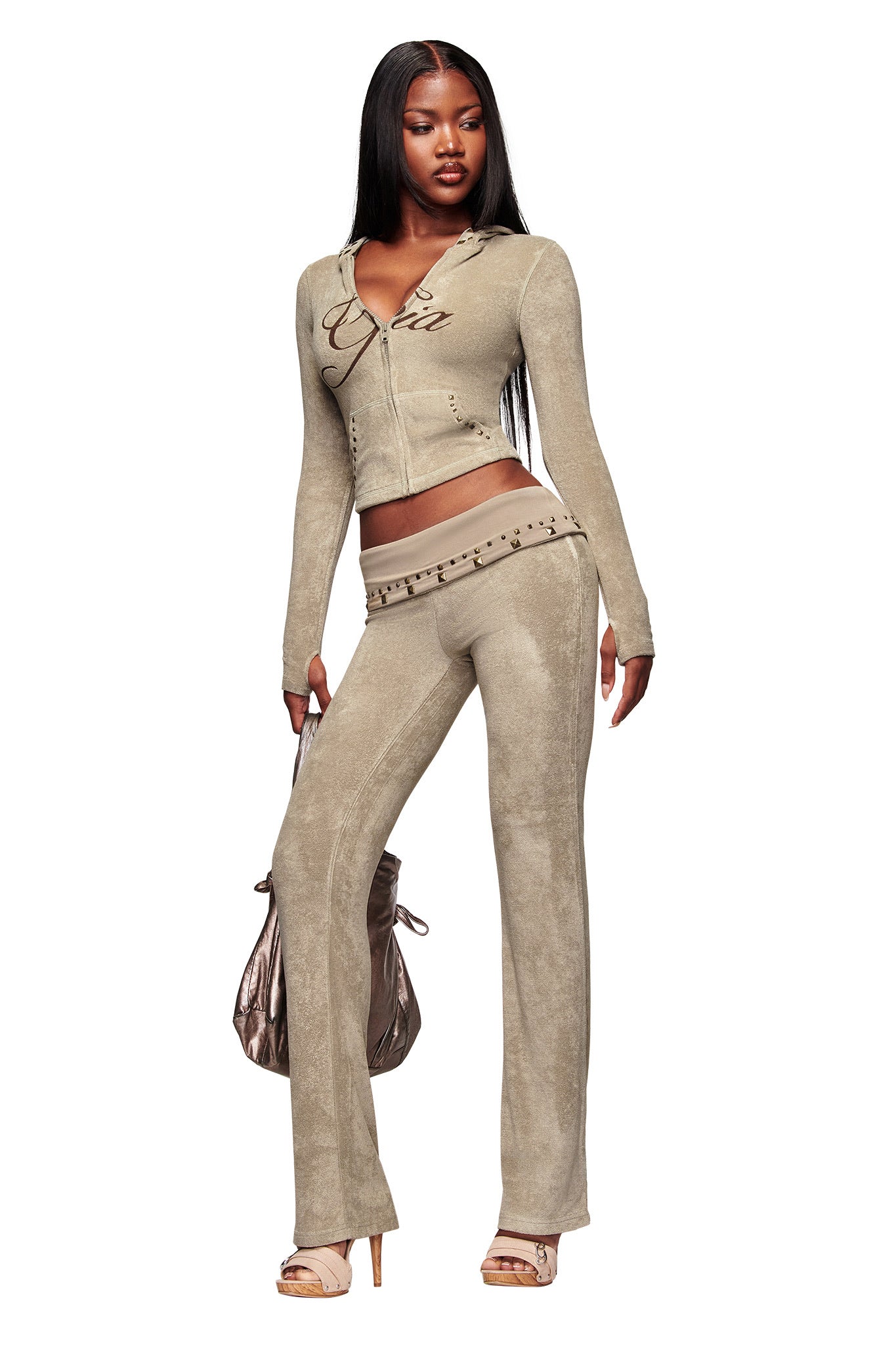 A woman confidently poses with a metallic handbag, wearing I.AM.GIA's BLARE STUDDED TERRY PANT in stone and a matching zip-up hoodie, paired with open-toe heels. She has long straight hair.