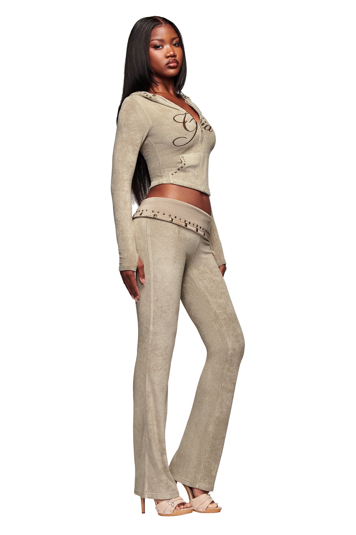 A woman stands in profile wearing I.AM.GIA’s BLARE STUDDED TERRY PANT in Stone with decorative stitching, a matching long-sleeve crop top, beige heeled sandals, long straight hair, and a confident expression.