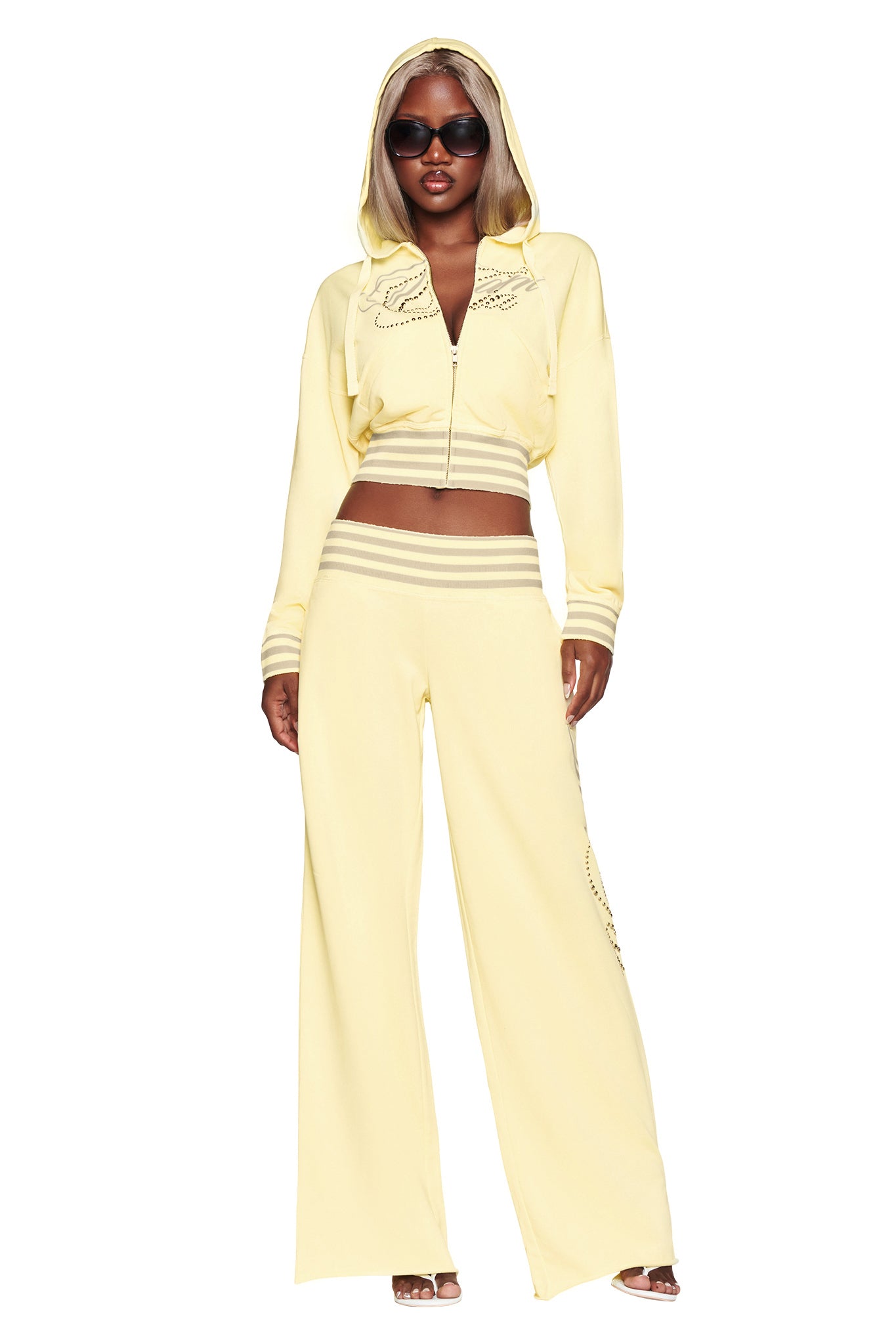 A woman wearing sunglasses and the I.AM.GIA VEDA PANT in yellow, paired with a light yellow hooded zip-up jacket and heels, stands facing forward against a white background.