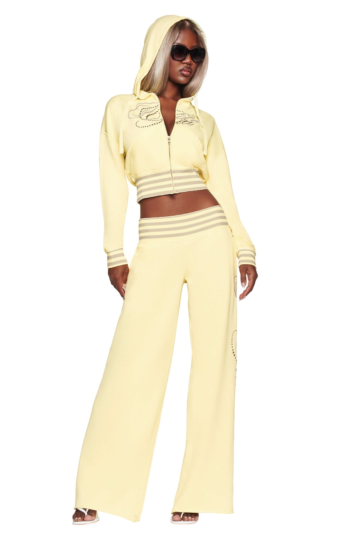 A woman stands confidently against a white background wearing the I.AM.GIA VEDA JACKET in yellow—a light yellow cropped zip-up hoodie with rhinestone script—paired with wide-leg pants, sunglasses, and open-toe heels.