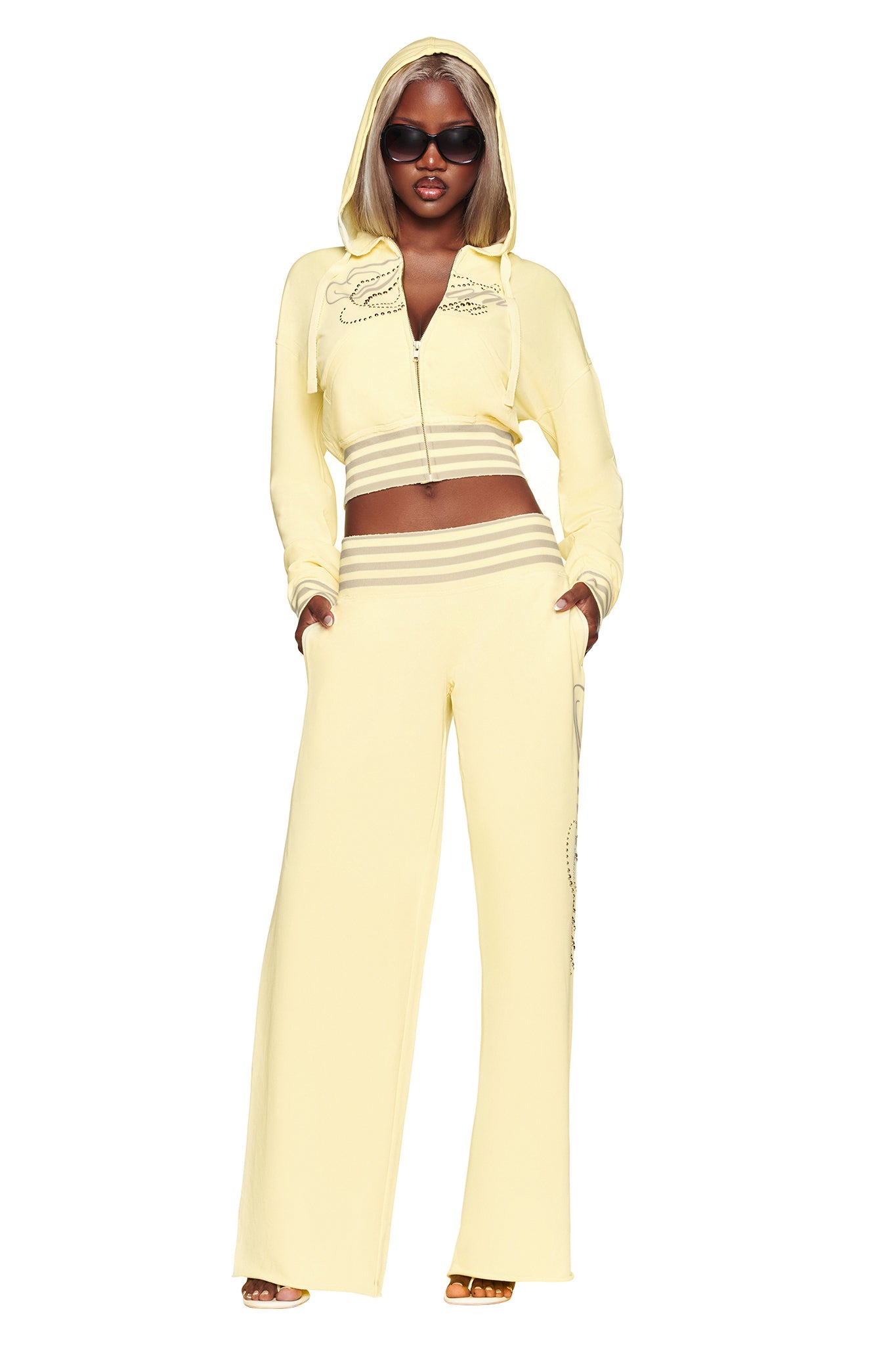 A woman wearing the I.AM.GIA VEDA PANT in yellow, styled with a matching yellow hooded zip-up crop top and sunglasses, stands with her hands in her pockets against a white background.