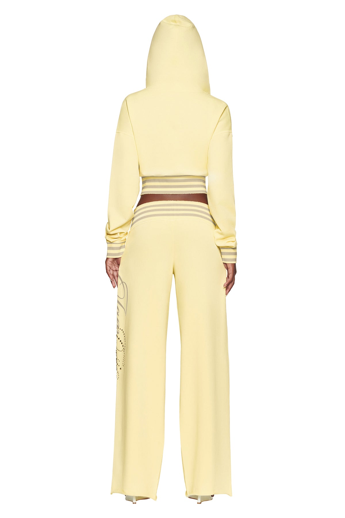 A person stands with their back to the camera, wearing I.AM.GIA’s VEDA PANT in yellow—a pale yellow, wide-leg pant featuring a hooded sweatshirt top, brown and white striped waistband, and a subtle design on the left pant leg.