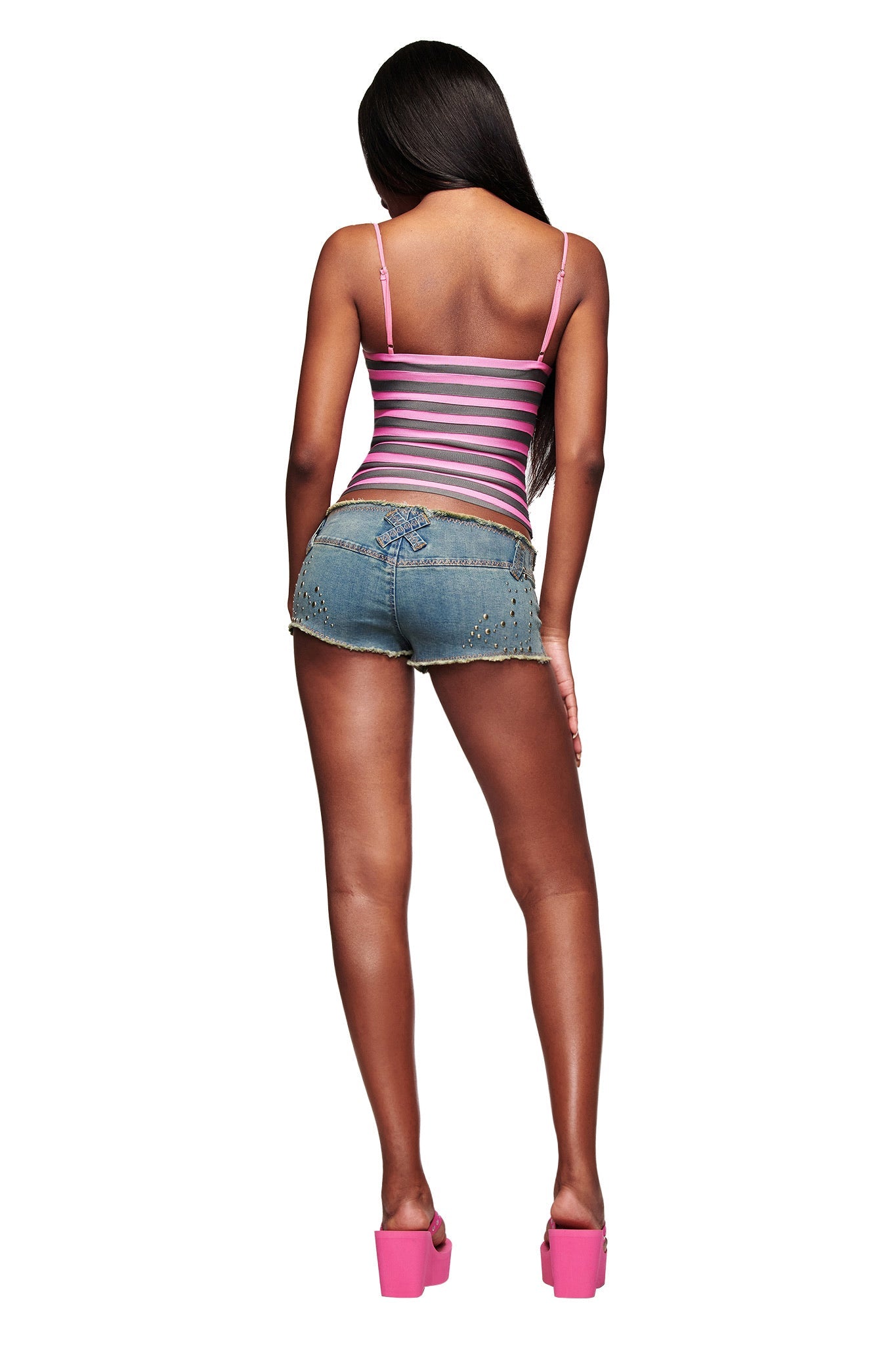 A woman with long hair faces away, wearing the I.AM.GIA NYNA BANDAGE CAMI TOP in hot pink, paired with denim shorts featuring studded details and pink platform heels, all set against a white background.
