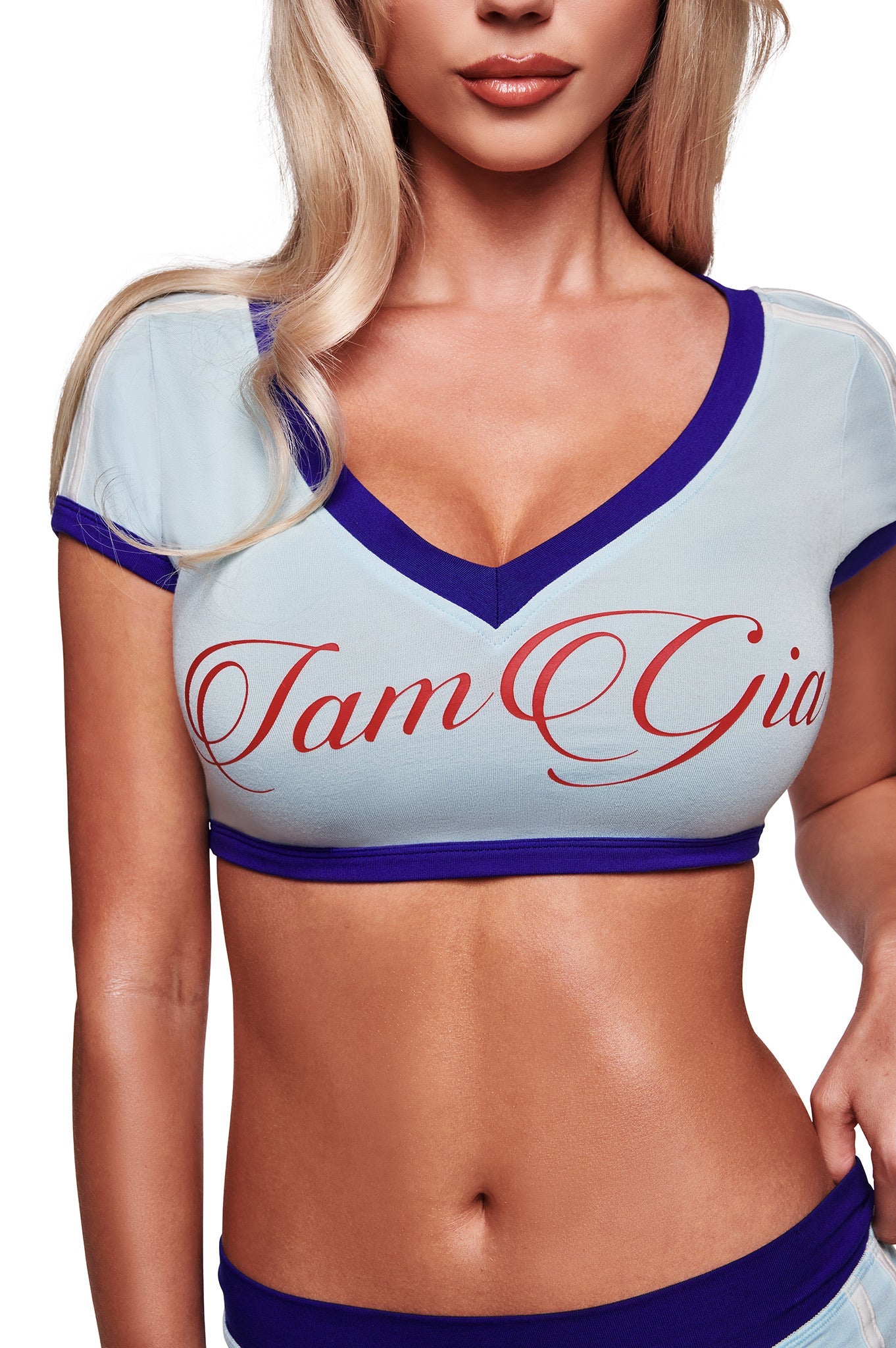 A woman with long blonde hair models the I.AM.GIA BLARE SPORT CROP TEE AND HEADBAND in baby blue, featuring dark blue trim and sporty v-neck styling. The image highlights the top’s modern design from chin to waist.