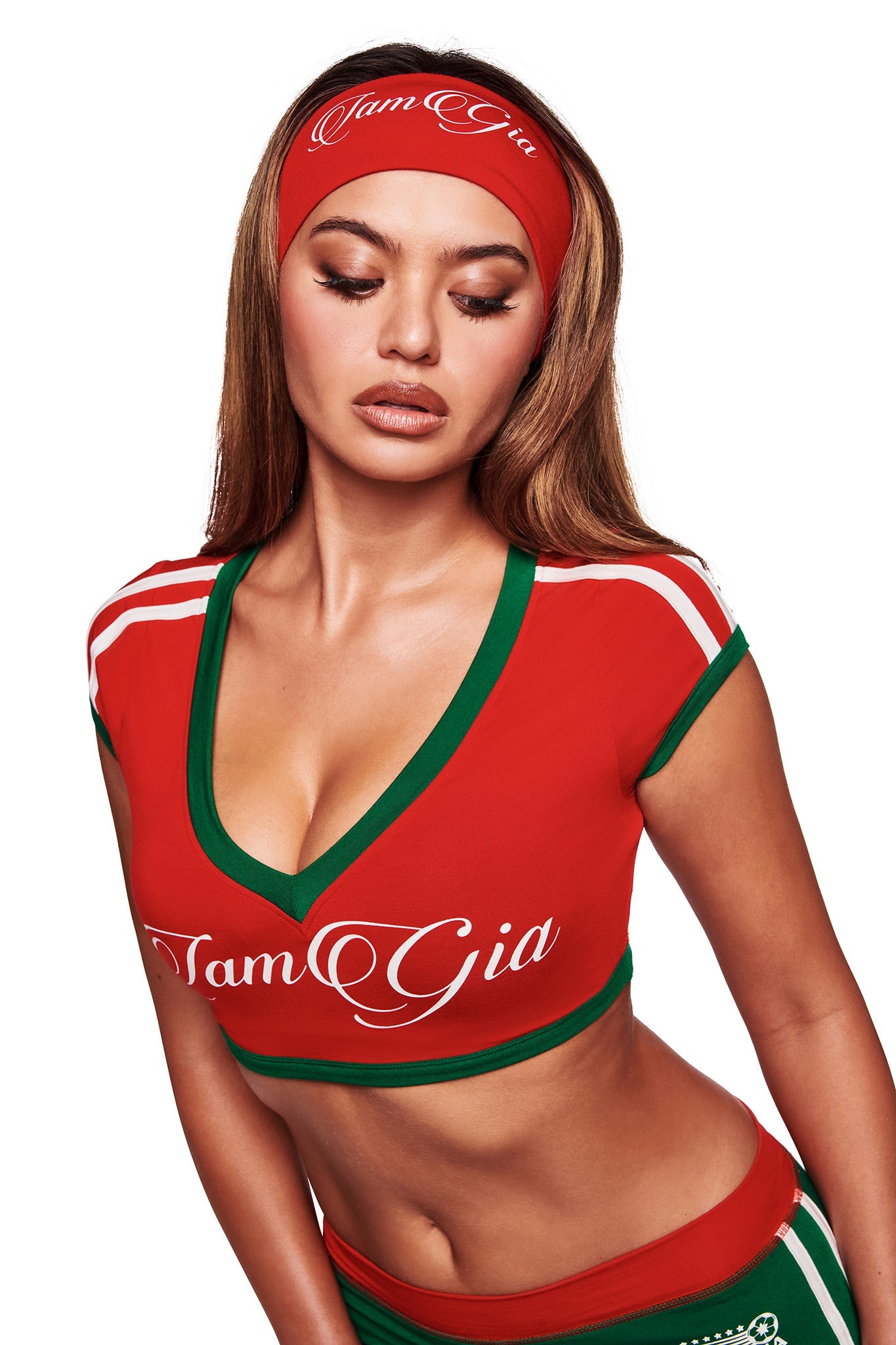 A woman with long hair wears the I.AM.GIA BLARE TWO-PIECE SPORT CROP TEE in crimson and a matching headband, both with green and white accents. She looks downward with a neutral expression against a white background.