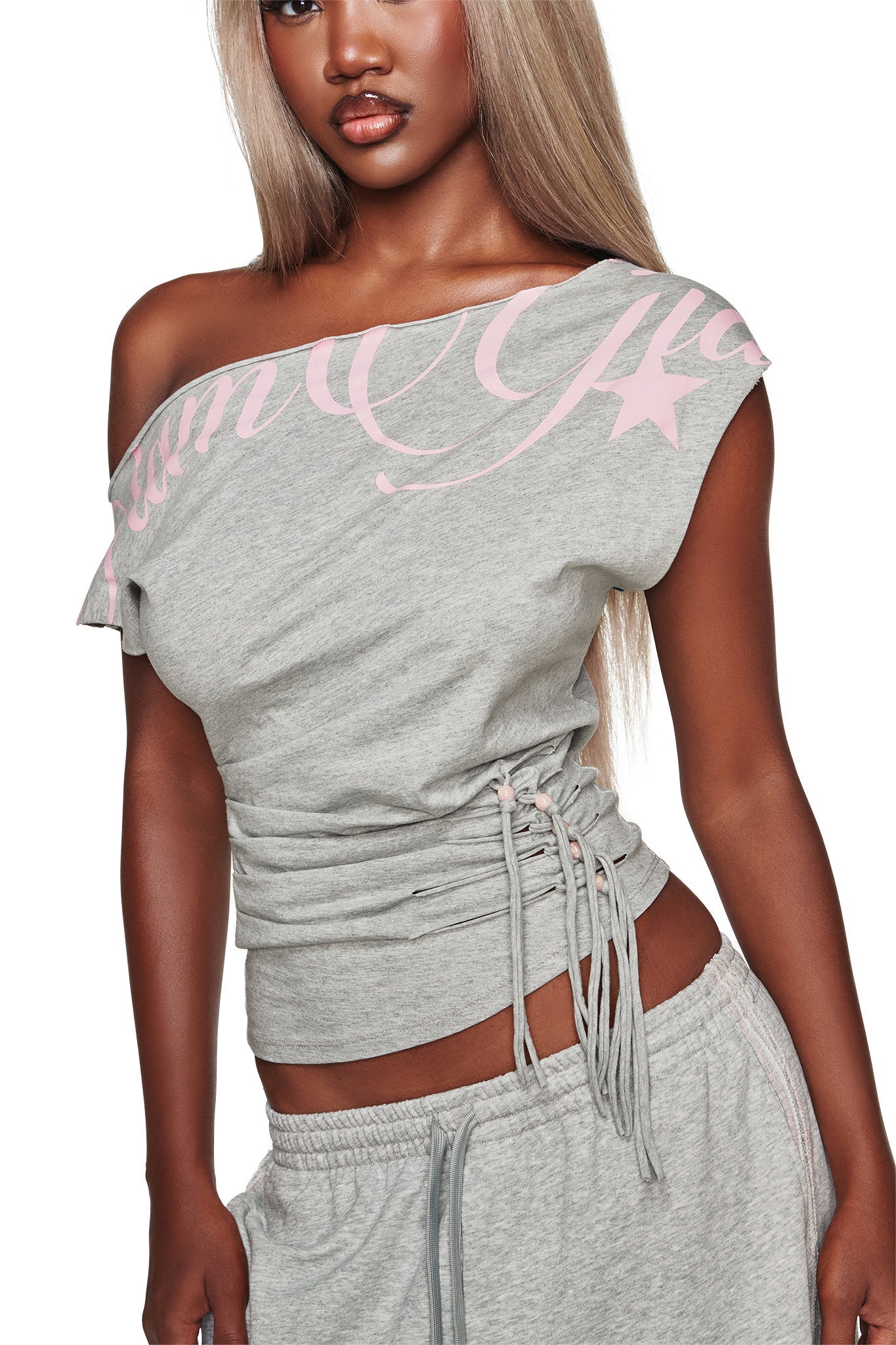 A woman with long blonde hair wears the BEX OFF SHOULDER TOP in grey by I.AM.GIA, featuring pink lettering. Only her upper body is visible against a white background.
