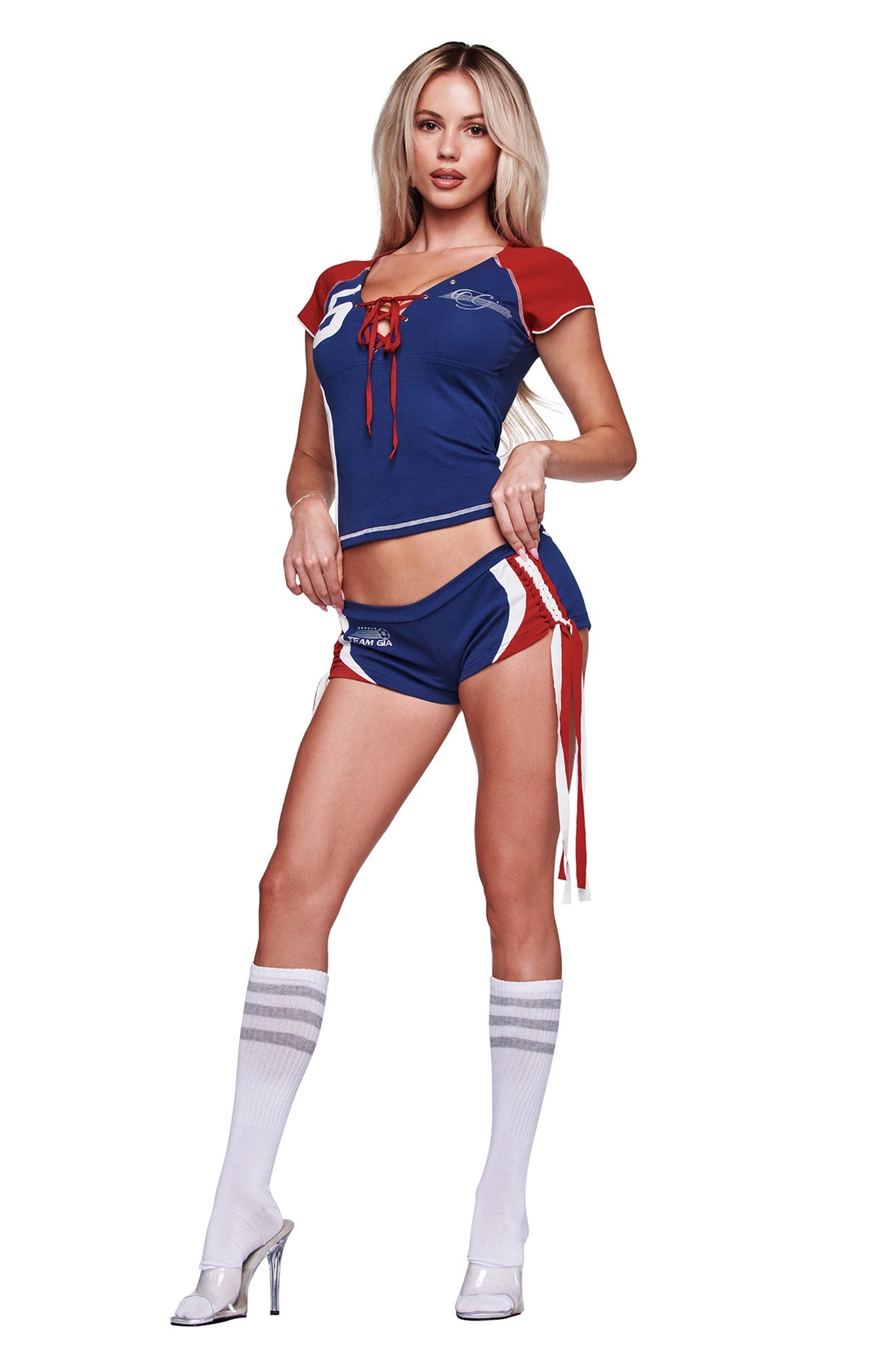 A woman poses against a white background wearing the I.AM.GIA NOVIA MICRO SPORT SHORT in navy, styled with knee-high white socks and high-heeled shoes for a sporty cheerleader-inspired look.