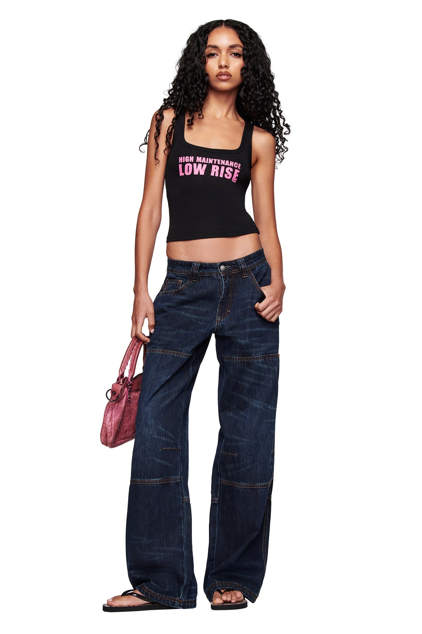 A woman with long curly hair wears a black "HIGH MAINTENANCE LOW RISE" crop top, I.AM.GIA MISTY JEAN - BLUE DUSTY WASH jeans, black flip-flops, and holds a pink handbag while standing against a white background.
