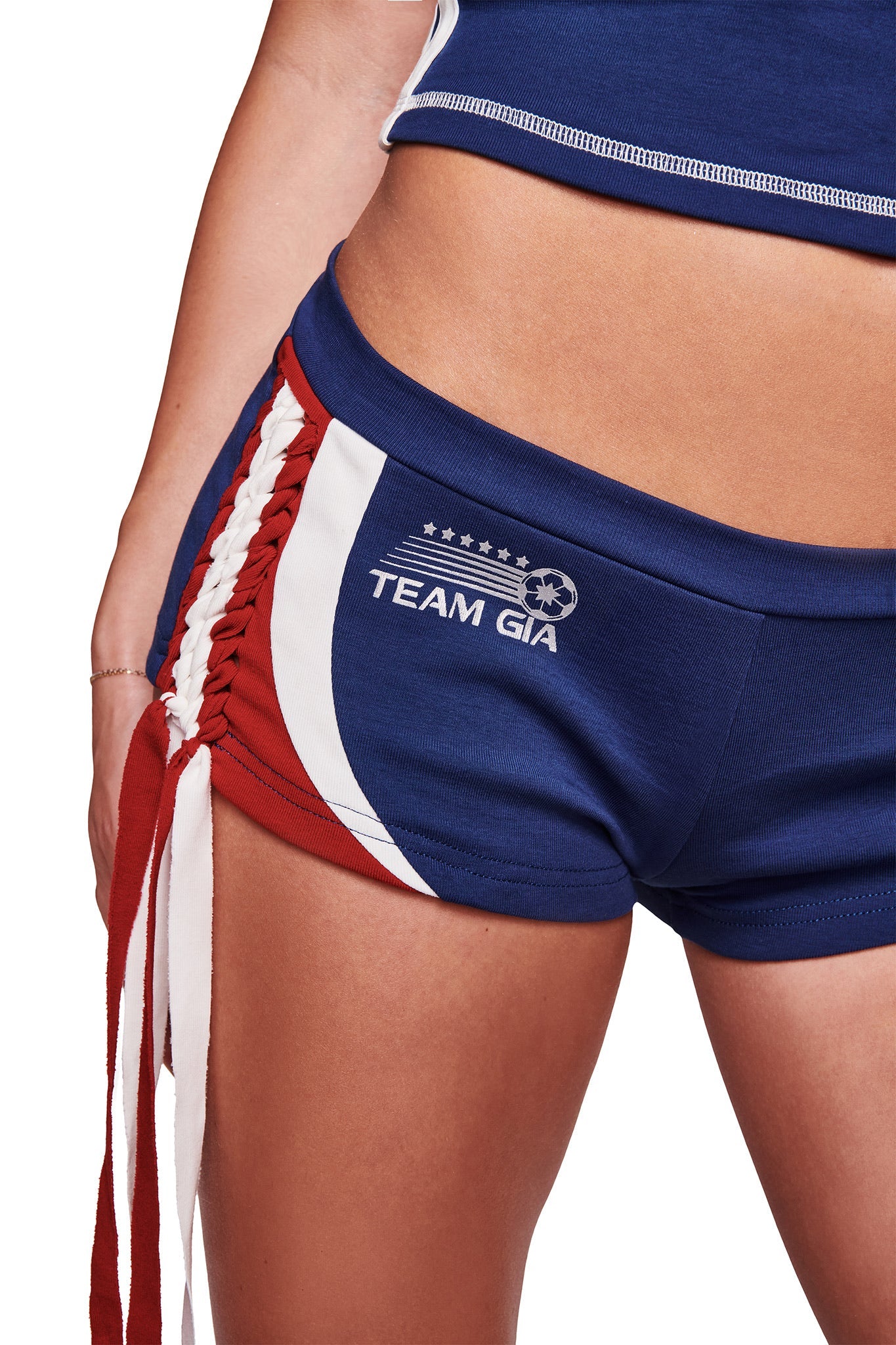 Someone wearing the I.AM.GIA NOVIA MICRO SPORT SHORT - NAVY: blue stretch cotton micro mini shorts with red and white side stripes, decorative side lacing, "TEAM GIA" on the left, and a low-rise fit. Only their upper body and legs are partly visible.