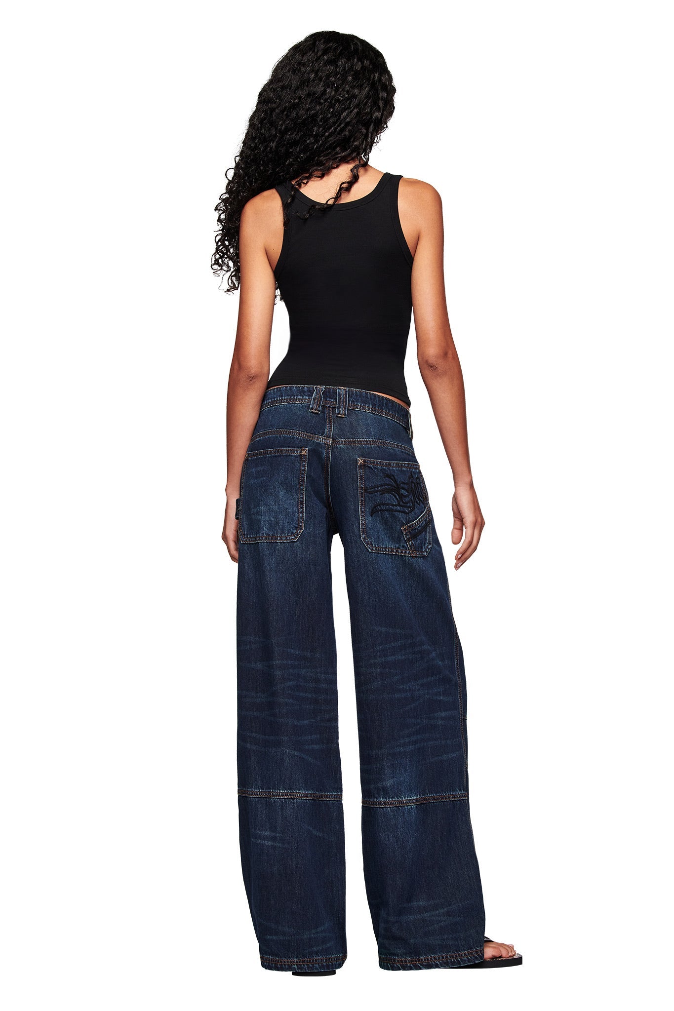 A woman with long curly hair wears the I.AM.GIA HOLLY TANK - HIGH MAINTENANCE and wide-leg dark blue jeans, standing with her back to a white background.