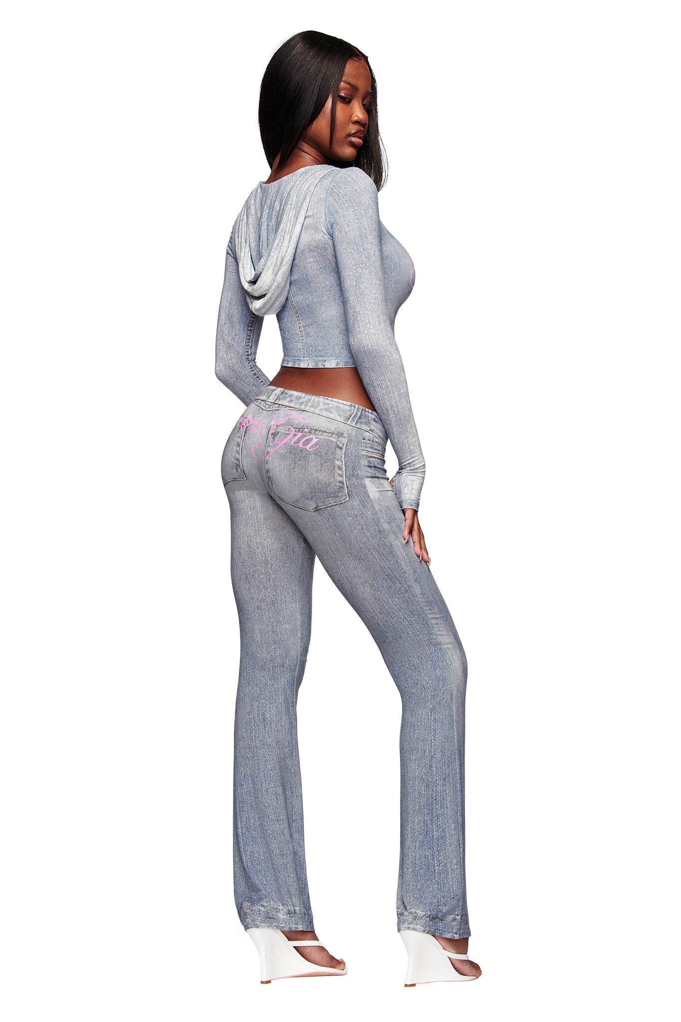 A woman models the I.AM.GIA BLARE TRACKPANT - DENIM TROMPE L'OEIL, a light blue faux-denim matching set with a long-sleeve hooded crop top and high-waisted pants featuring pink writing on the back, styled with white open-toe heels.