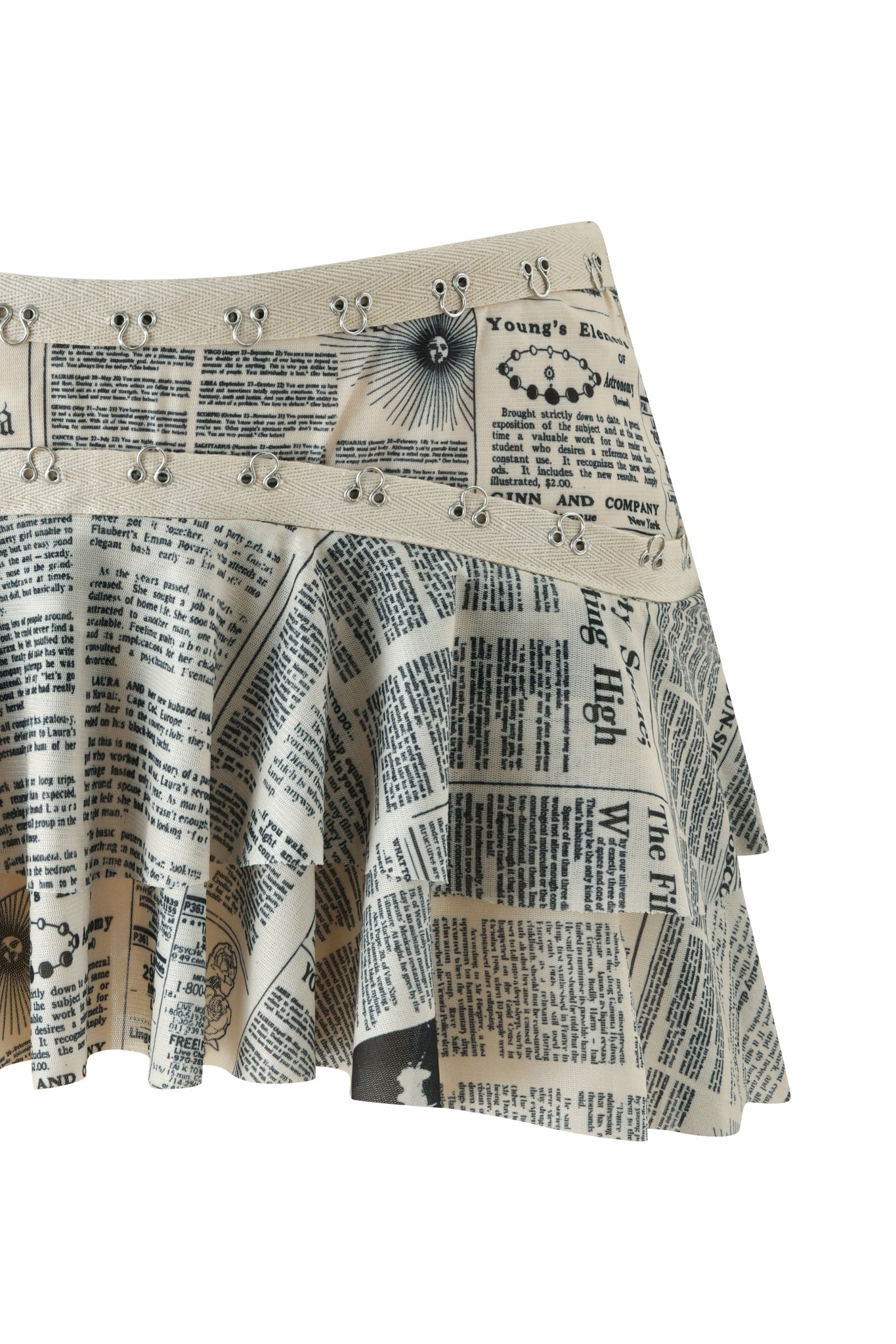 The SORA MINI SKIRT - LEFT ON READ by I.AM.GIA is a pleated, low-rise mini skirt featuring a striking black and white newspaper print with bold text, graphic elements, and edgy metal grommet details along the waistband.