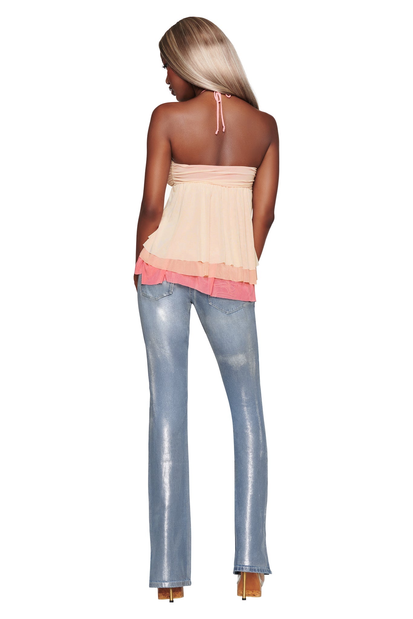 A woman with long blonde hair stands with her back to the camera, wearing the I.AM.GIA IDALIA TOP in Raspberry Multi—a mesh halter neck style adorned with cascading ruffles in peach and pink tones—paired with shiny light blue jeans and high heels.