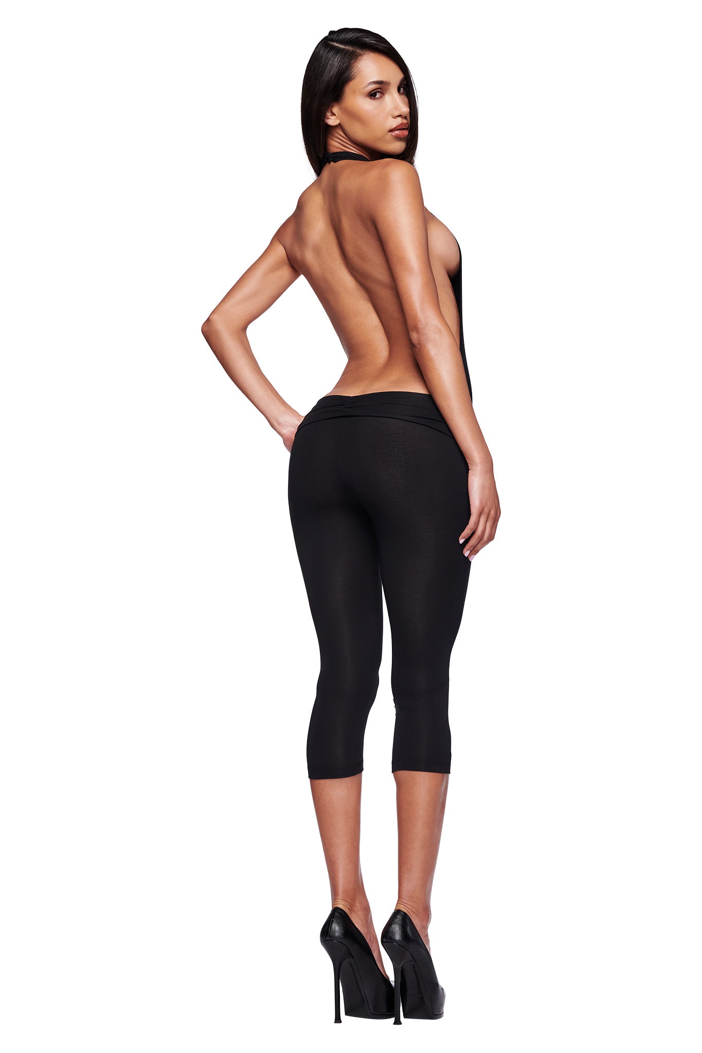 A woman models the I.AM.GIA KAIRA JUMPSUIT - BLACK, featuring a ruched waistband, paired with high heels. She stands with her back to the camera, looking over her shoulder against a white background.