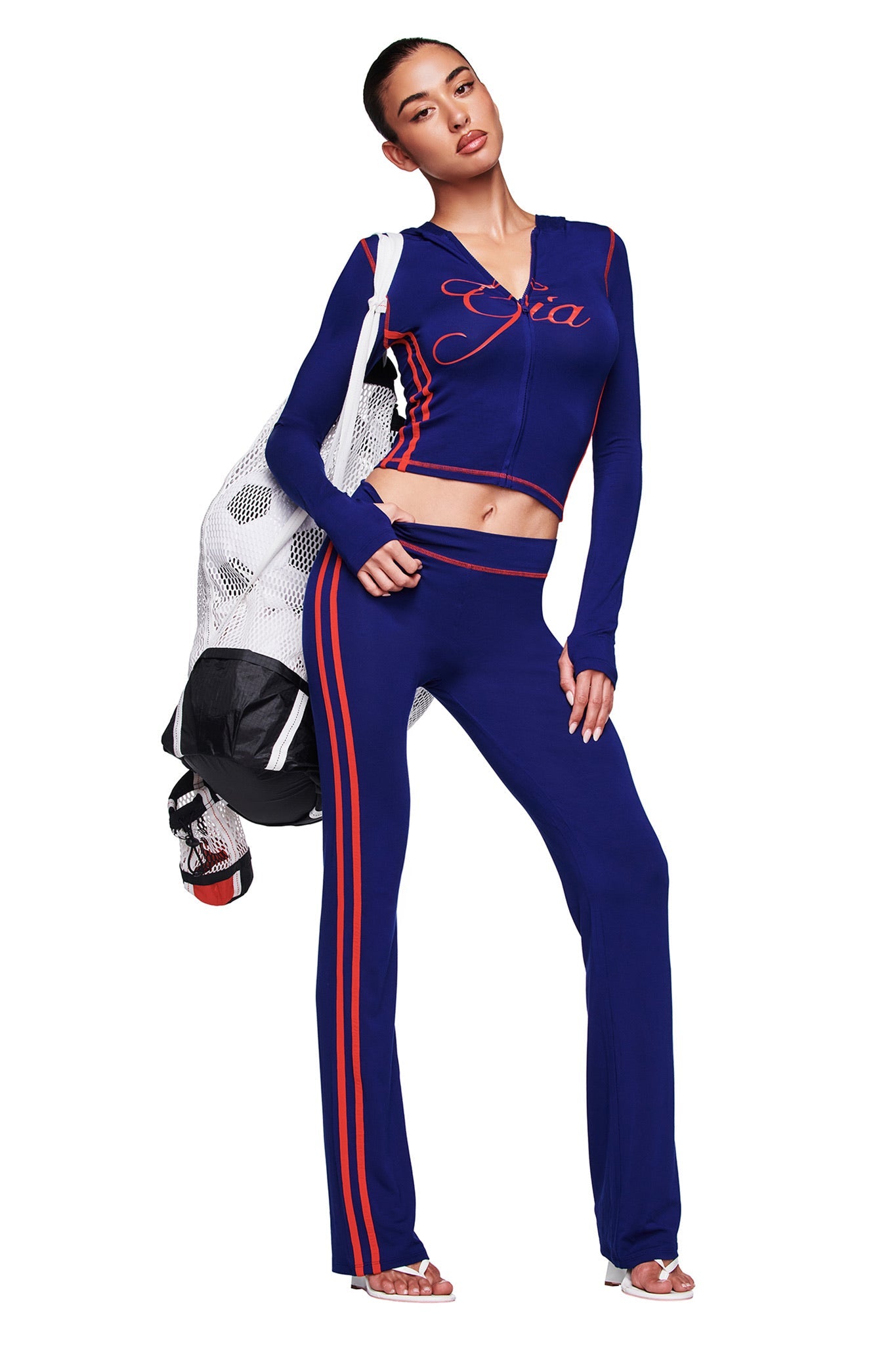 A woman confidently poses in I.AM.GIA's BLARE SPORT TRACKPANT - NAVY and a blue "Gia" top, holding a white mesh bag with black and red items against a white backdrop. The buttery-soft fabric brings comfort to her bold style.
