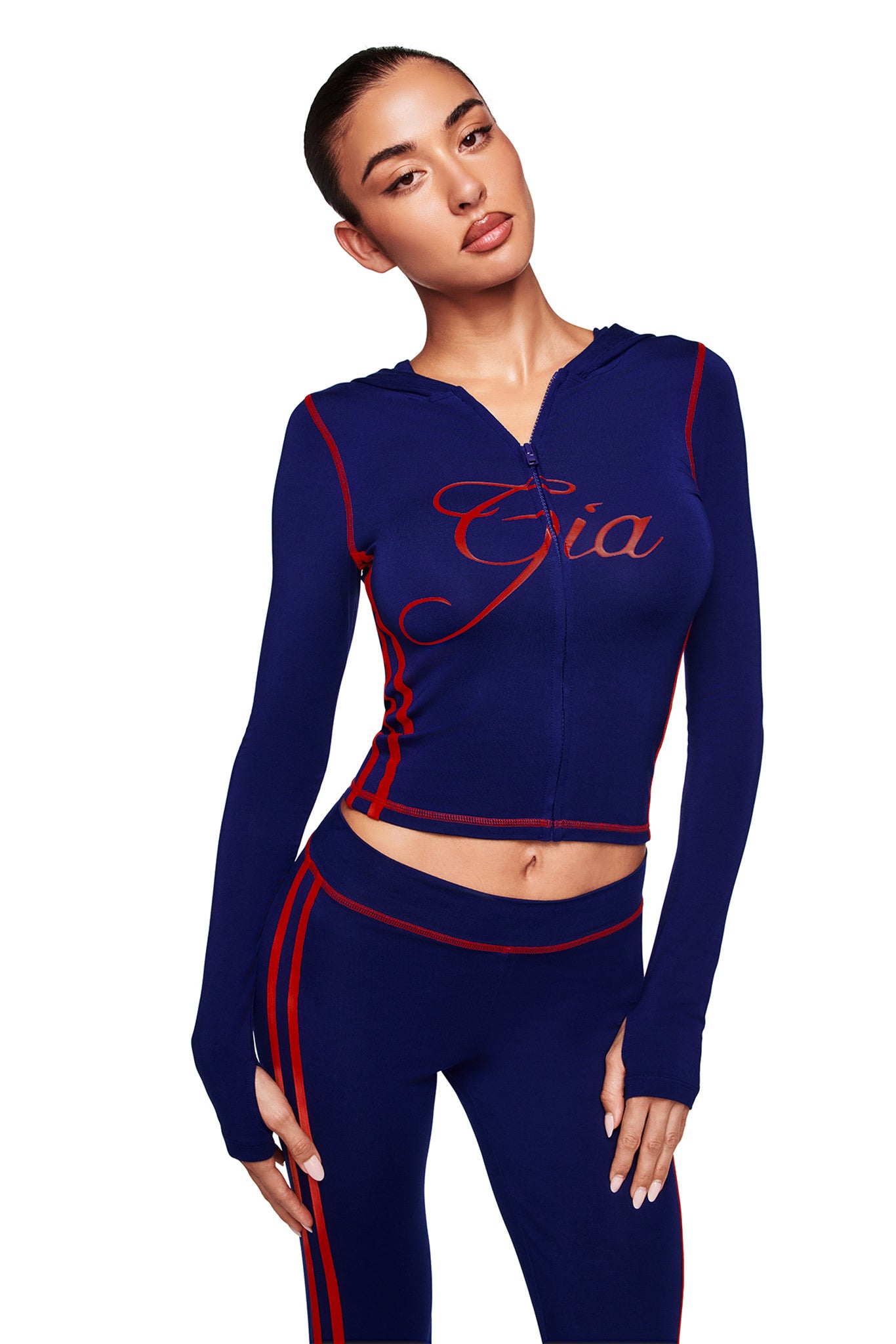 A woman models the I.AM.GIA BLARE SPORT HOODIE AND HEADBAND in navy, featuring red trim, buttery-soft fabric, and a fitted cropped zip-up design, against a white background.