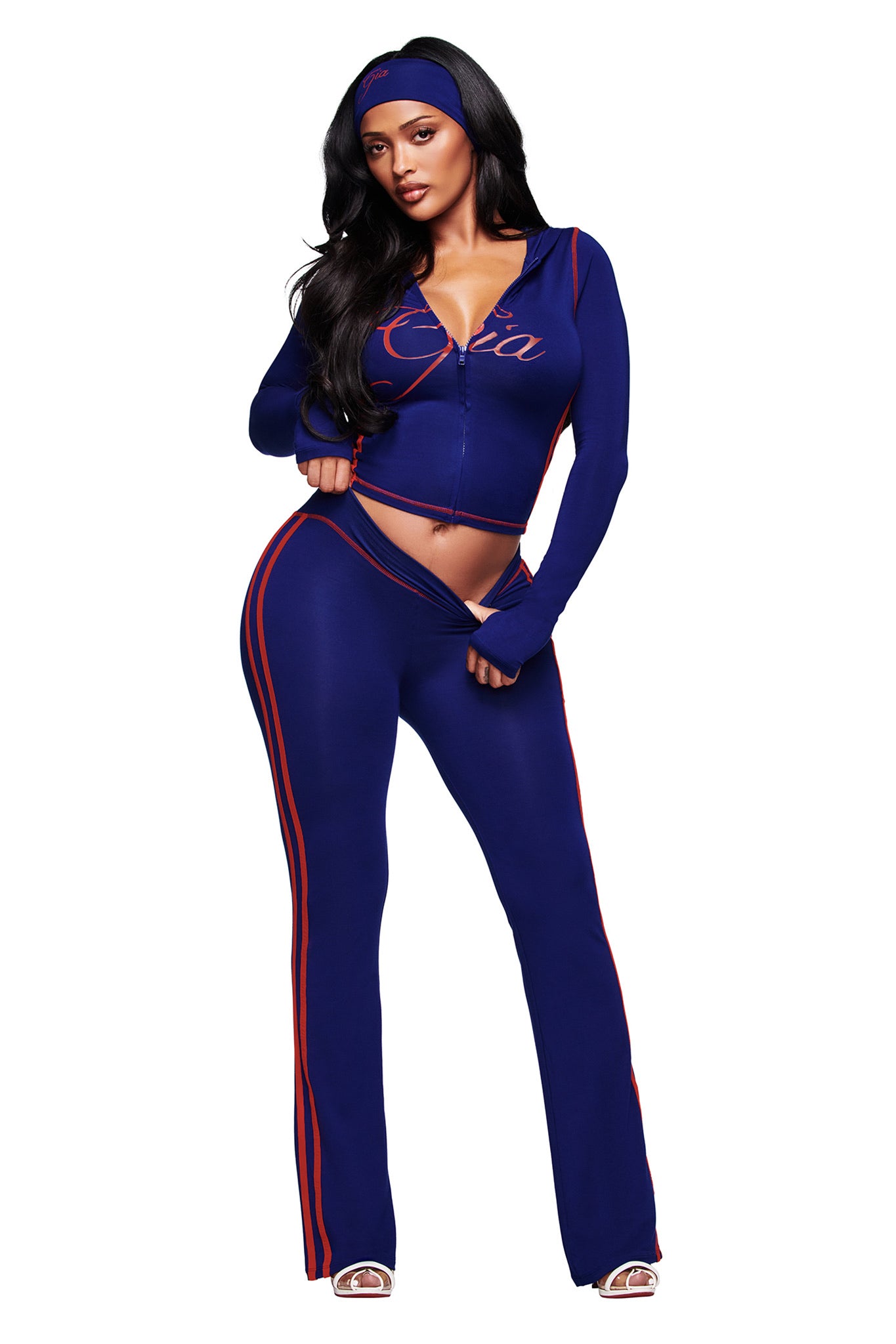 A woman confidently wears the I.AM.GIA BLARE SPORT HOODIE AND HEADBAND in navy, featuring buttery-soft fabric and a zip-up top, pairing effortlessly with open-toe heels for a bold, stylish look.