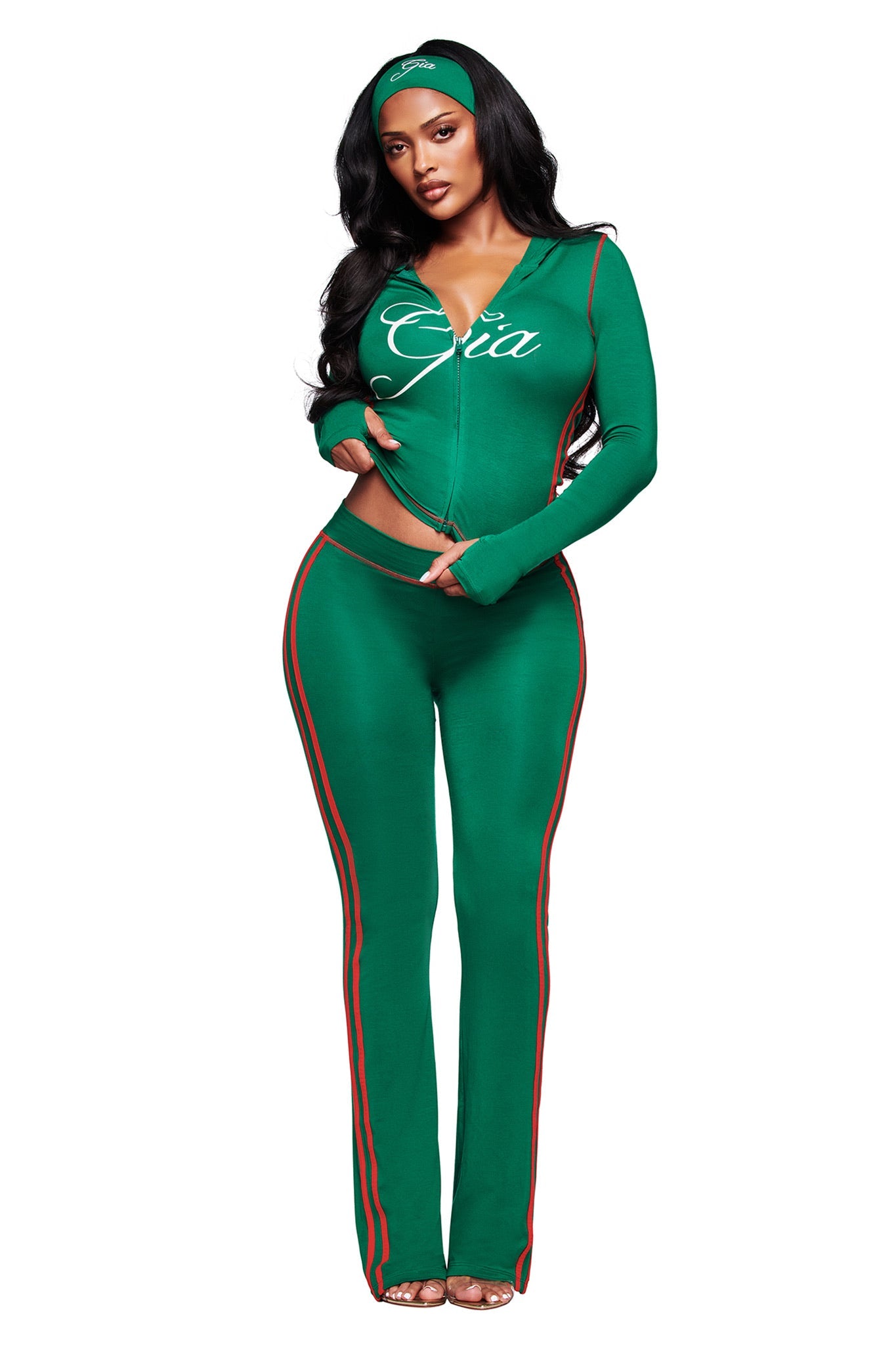 A woman wears the I.AM.GIA BLARE TWO-PIECE SPORT HOODIE in green with red and white stripes, featuring a fitted zip-up top reading "Gia," a matching headband, and open-toe heels, standing against a white background.