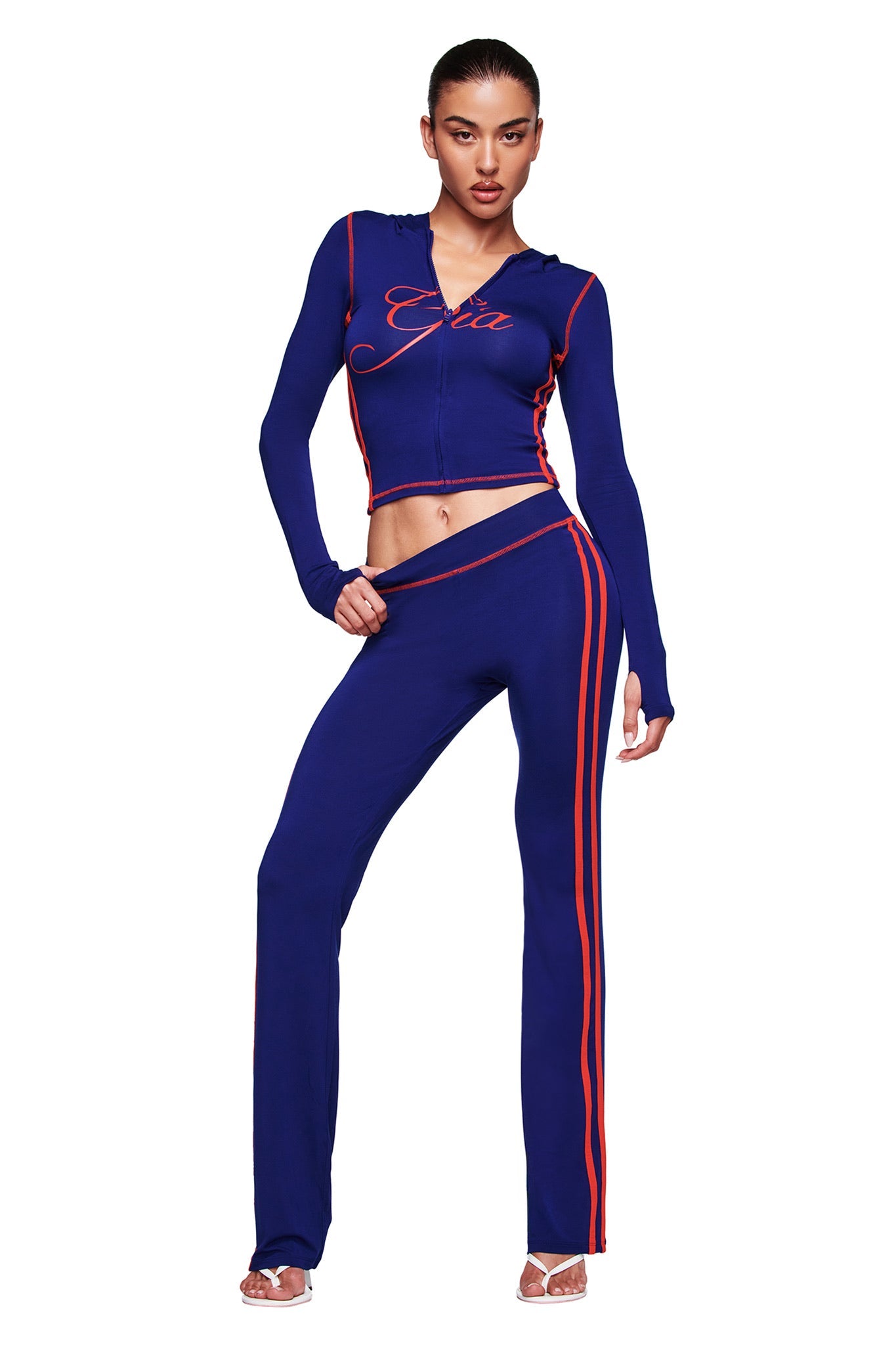A woman models the I.AM.GIA BLARE SPORT TRACKPANT in navy, featuring buttery-soft fabric and red side stripes, paired with a matching zip jacket with "Gia" on the chest. She wears clear heels against a white backdrop.