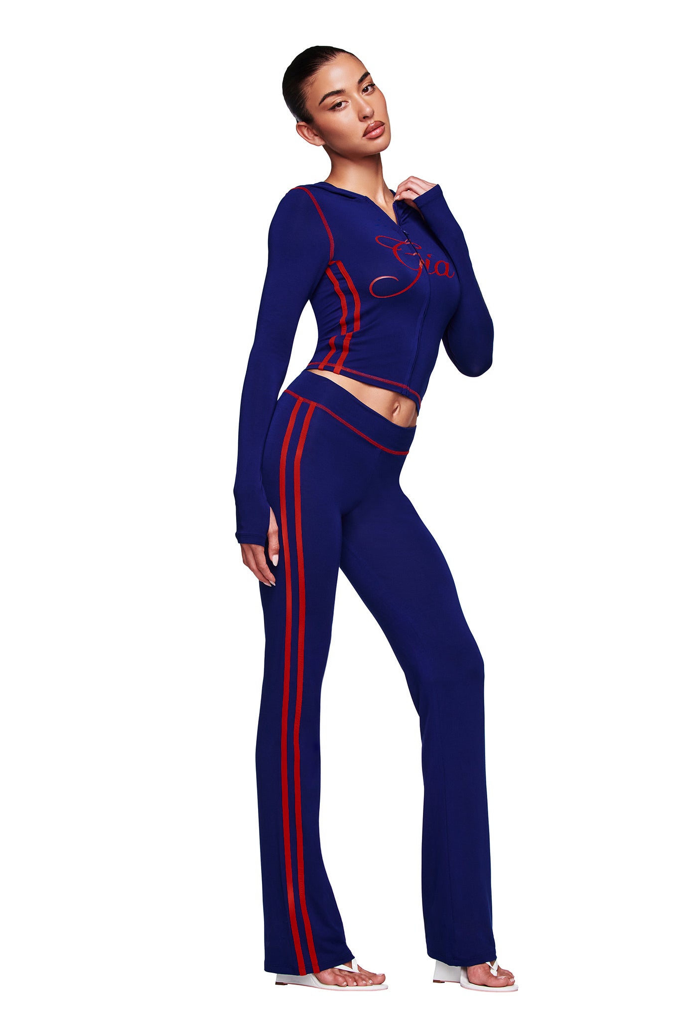 A woman poses in the I.AM.GIA BLARE SPORT TRACKPANT - NAVY, a fitted two-piece with red stripes and lettering, crafted from buttery-soft fabric. The set includes long flared pants and a long-sleeved top. She stands in open-toe heels against a white background.