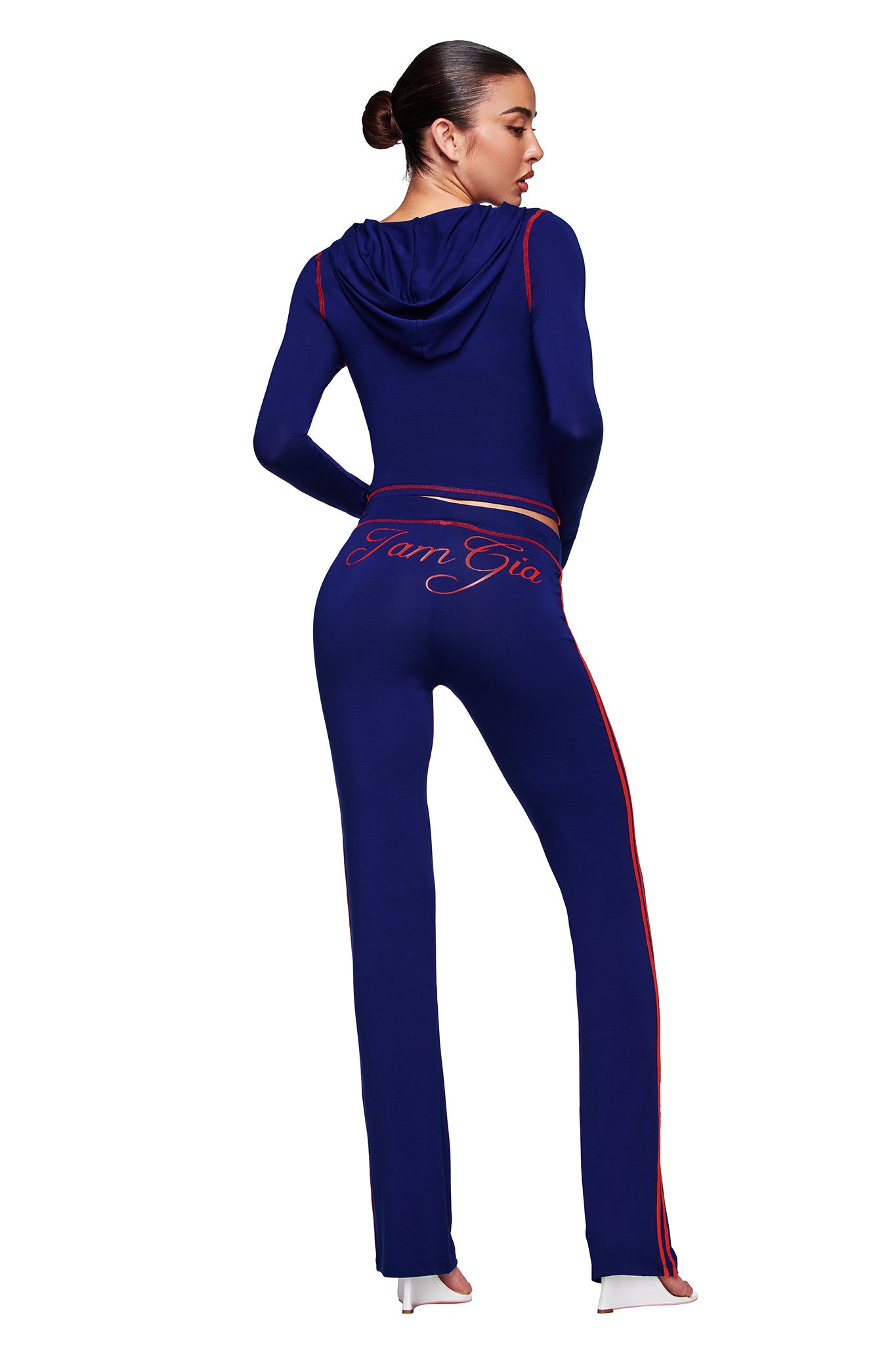 A woman faces away wearing the I.AM.GIA BLARE SPORT HOODIE AND HEADBAND in navy, featuring red piping, a hood, thumb-hole detail, and "I.AM.GIA" in cursive on the back of the pants. She stands in high heels against a white background.