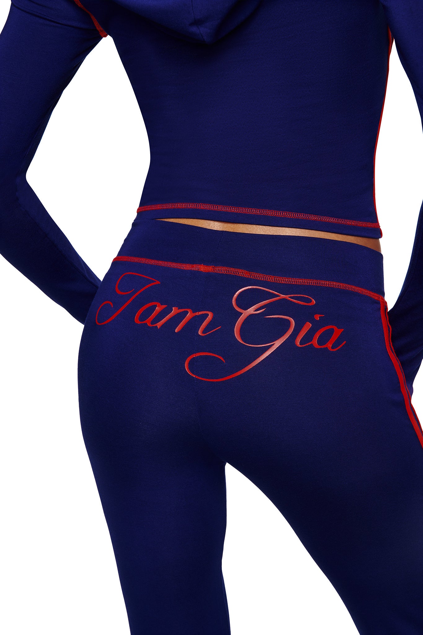 A person wears the I.AM.GIA BLARE SPORT TRACKPANT in navy with red trim and large cursive "I.AM.GIA" lettering across the back. The image is cropped to show only their torso and upper legs.