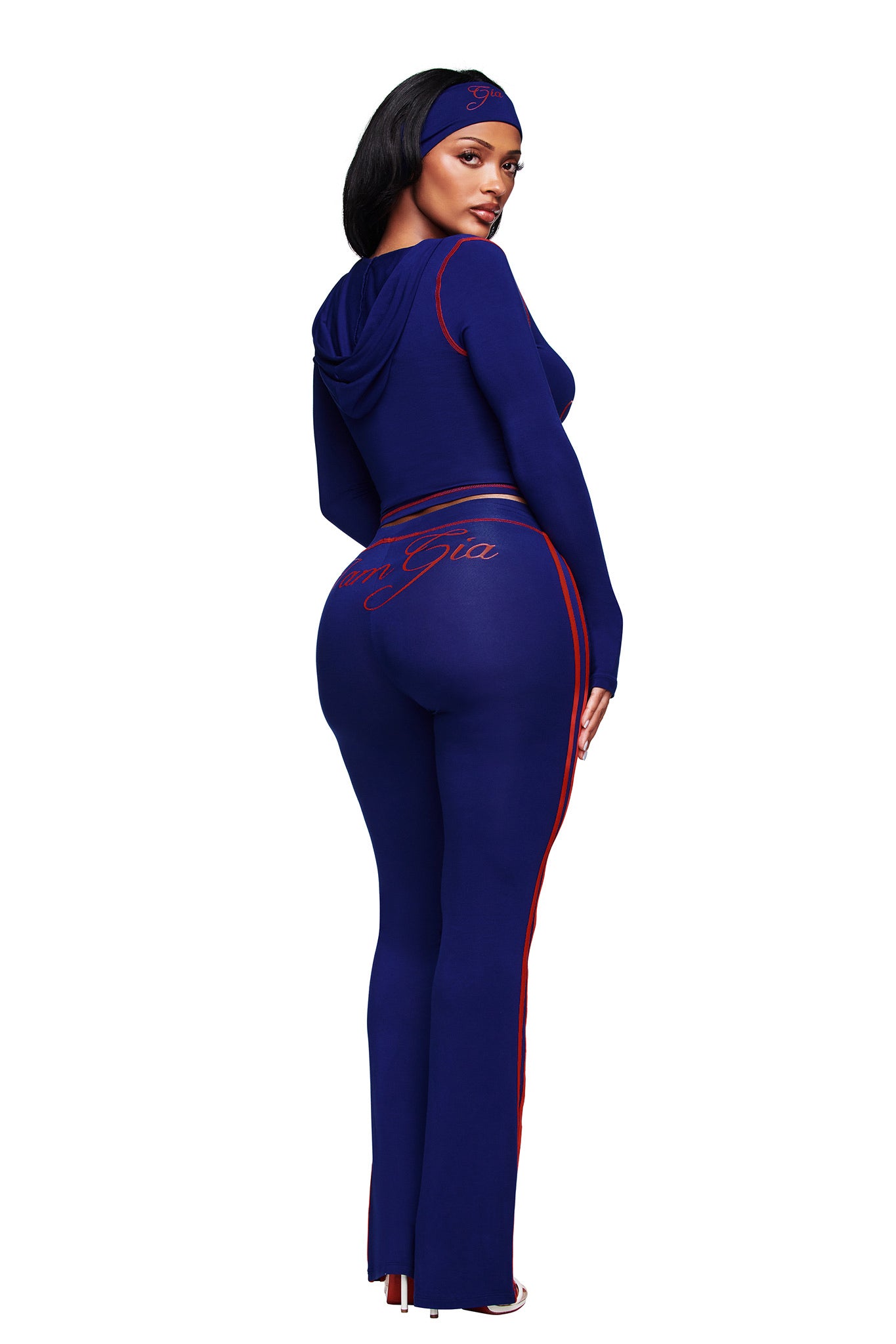 A woman faces away, wearing the I.AM.GIA BLARE SPORT TRACKPANT in navy with red stripes, hood, and matching headband. Cursive writing appears on the back of her pants as she looks over her shoulder against a white background.
