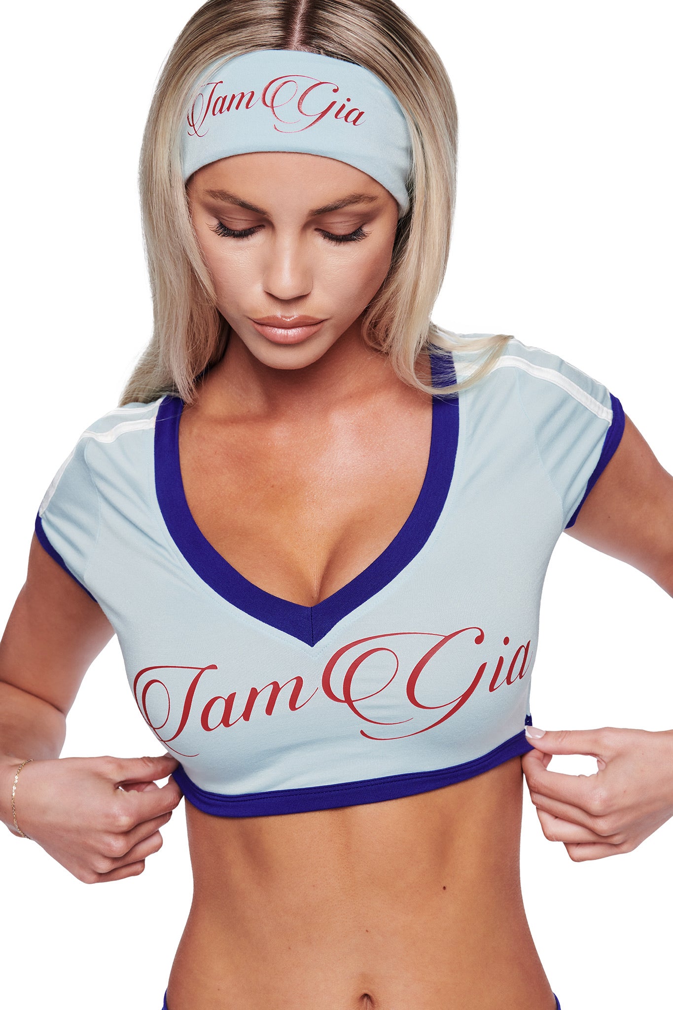 A woman with long blonde hair wears a blue crop top with "I am Gia" in red script and an I.AM.GIA BLARE SPORT HEADBAND in baby blue, looking down as she adjusts her shirt.