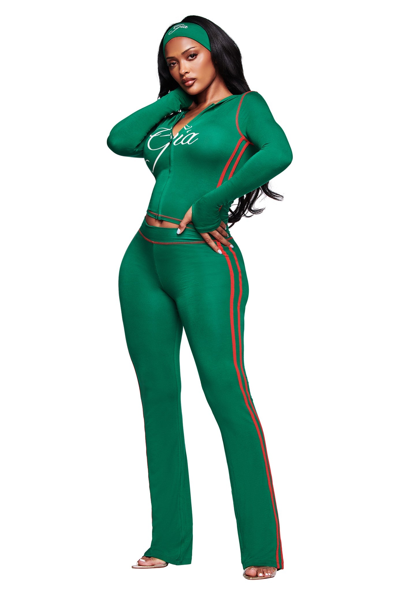 A woman poses confidently in a fitted green tracksuit with red and orange side stripes. She wears the I.AM.GIA BLARE SPORT HEADBAND - GREEN, while her jacket features "Ciao" on the chest, and her long dark hair falls in loose waves.