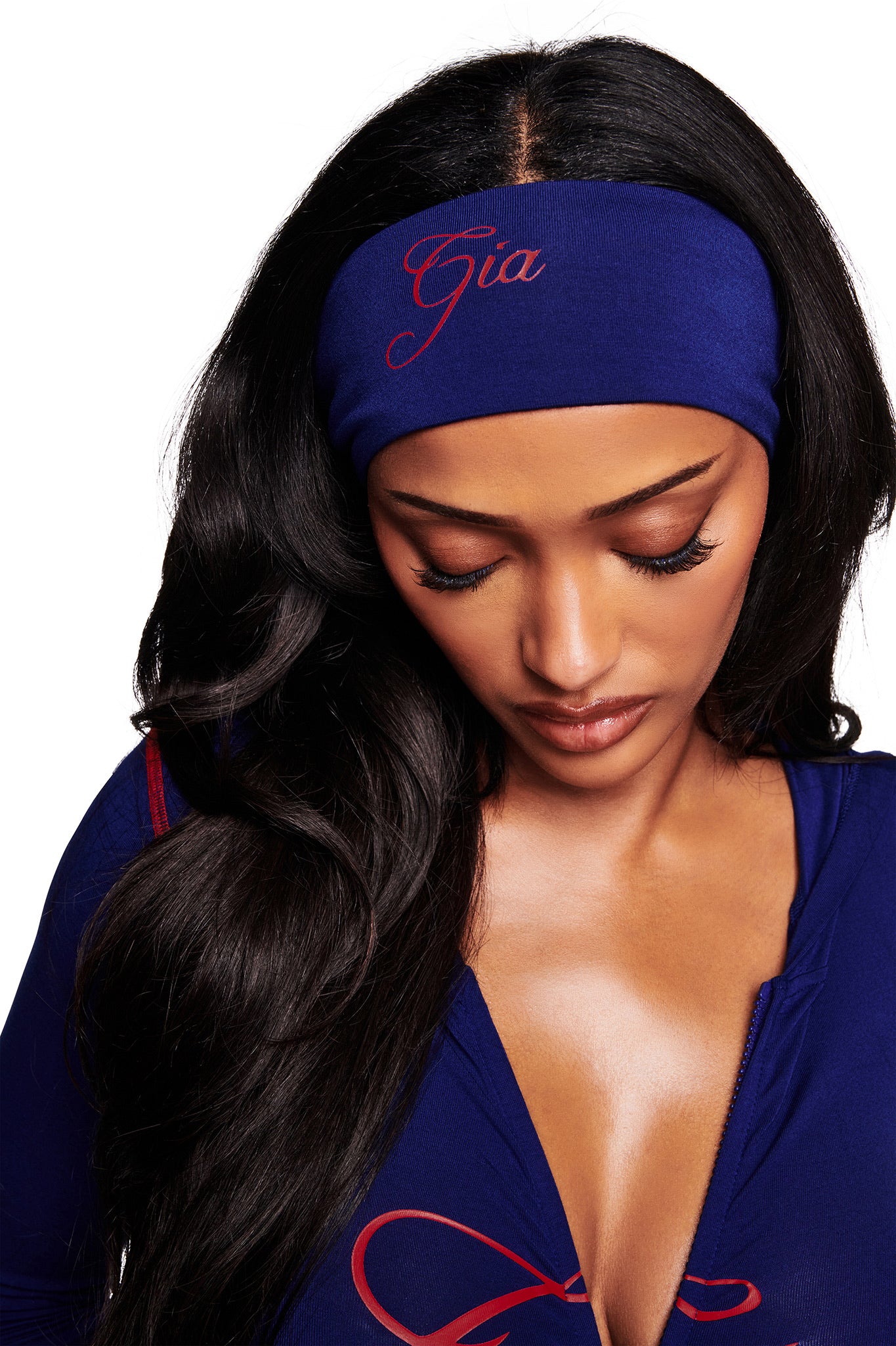 A woman with long, dark hair looks down, wearing the I.AM.GIA BLARE SPORT HEADBAND in navy, featuring red "Gia" embroidery. She pairs it with a deep-neck blue top against a plain white background.