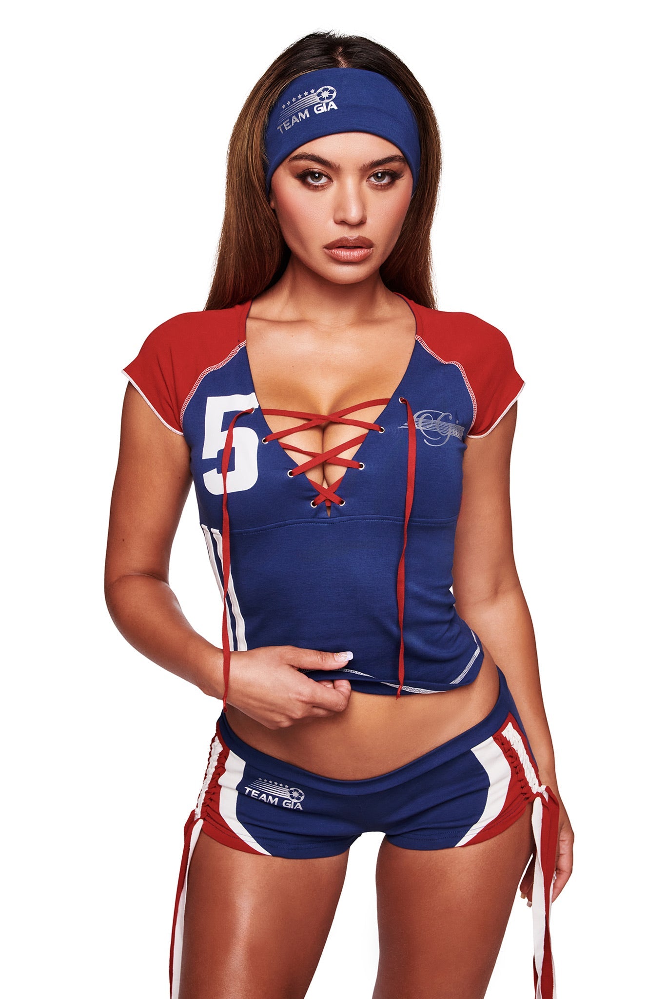 A woman poses with one hand on her waist in a blue and red sporty outfit, featuring a lace-up front top, matching shorts, and the I.AM.GIA NOVIA SPORT HEADBAND in navy, against a white background.