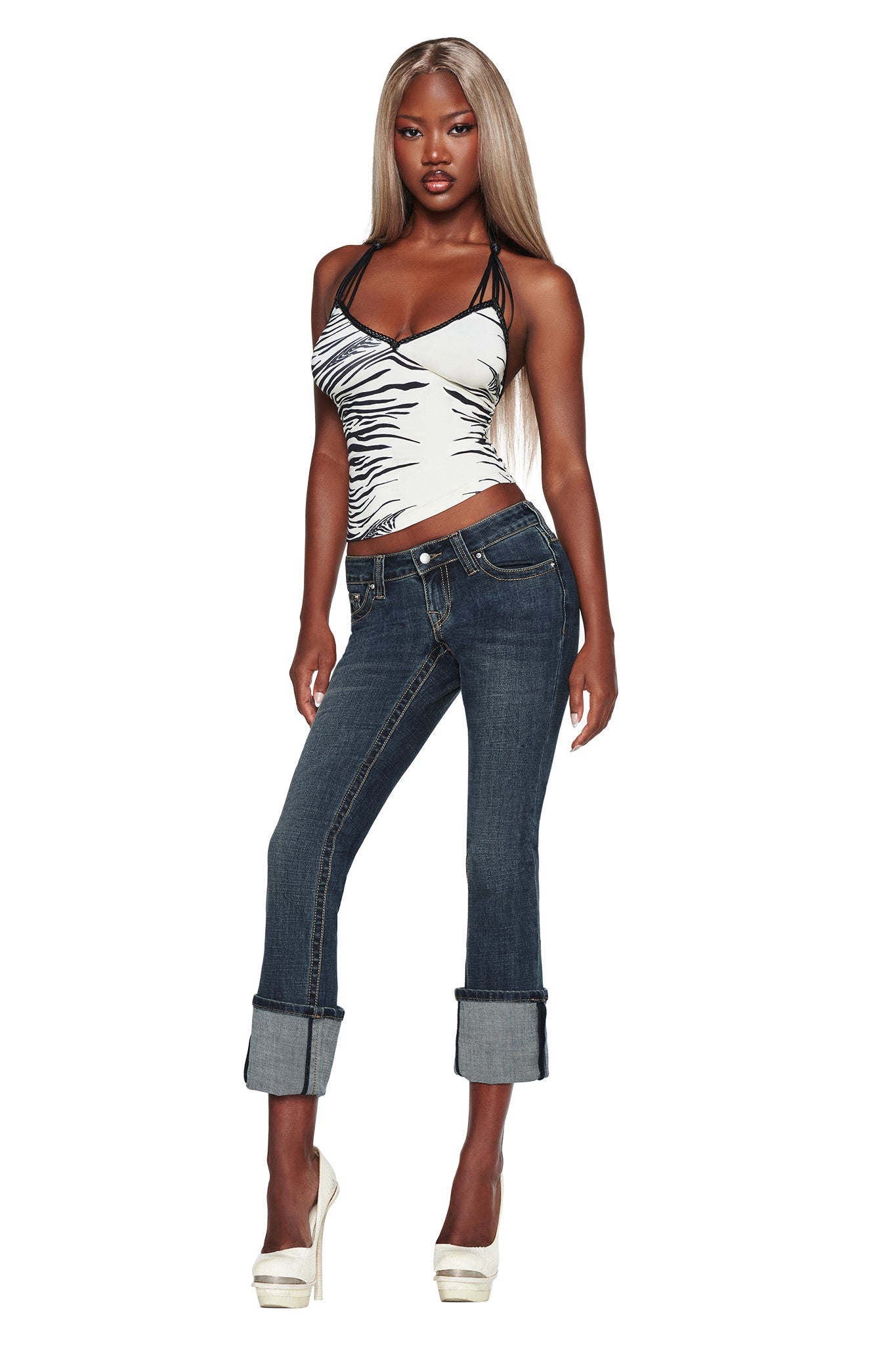 A woman with long, straight blonde hair strikes a confident pose against a white background, wearing the I.AM.GIA BIANKA JEAN - BLUE in a slim straight-leg, low-rise fit. She pairs the jeans with a black-and-white halter top and white high heels.