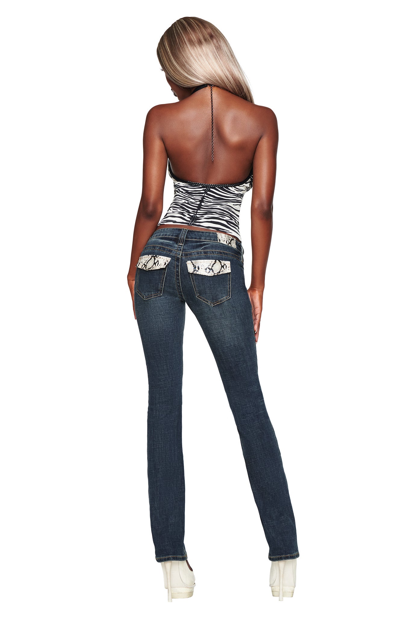 A woman stands with her back to the camera, wearing the I.AM.GIA BIANKA JEAN - BLUE in a low-rise, slim straight-leg fit with patterned pockets, paired with a zebra-print halter top and high platform heels against a white background.