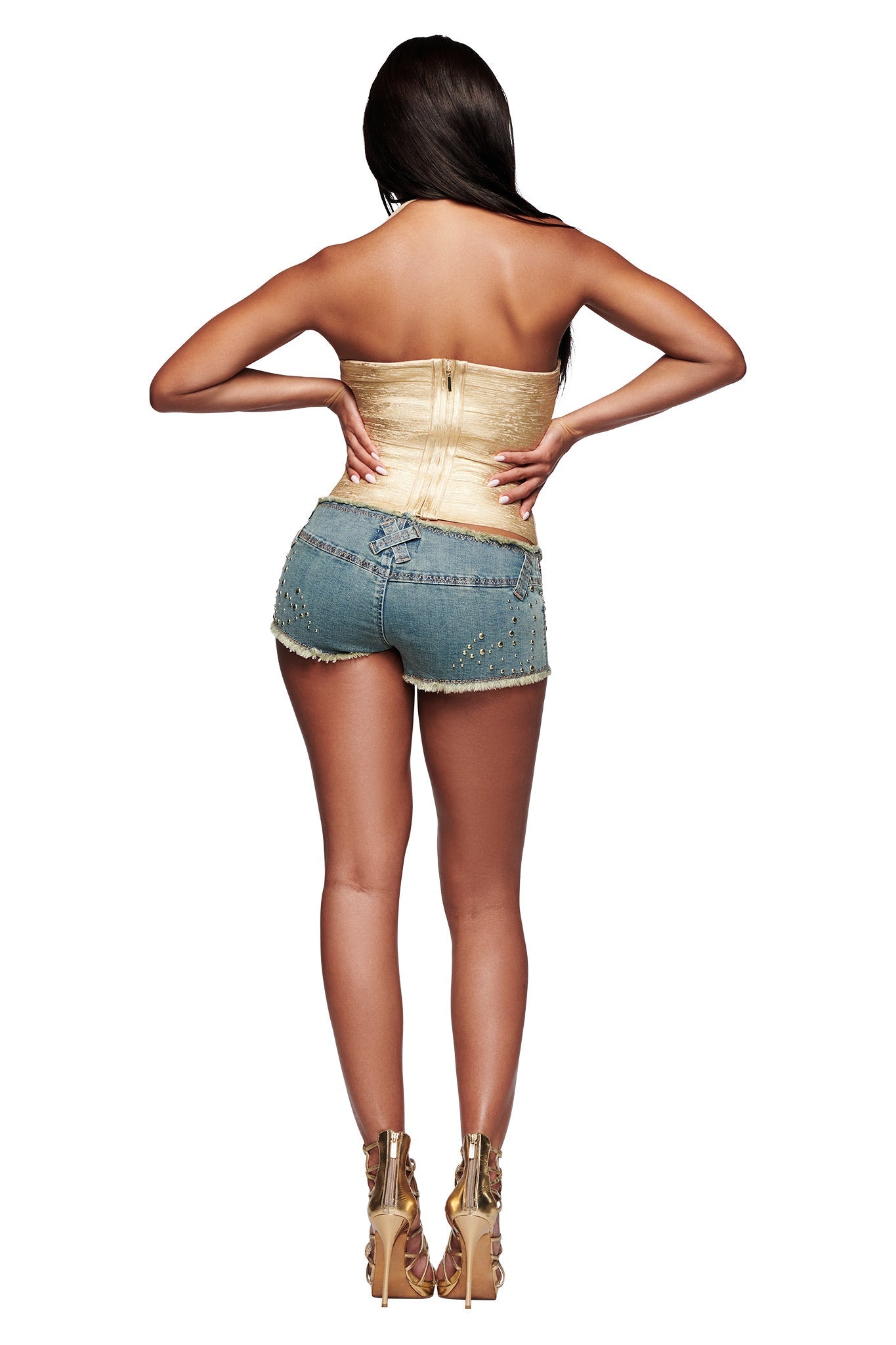 A woman faces away from the camera, hands on hips, wearing the I.AM.GIA AIDEN PLATINUM BANDAGE TOP in gold, paired with studded denim shorts and gold heels. Her long hair cascades down her back.