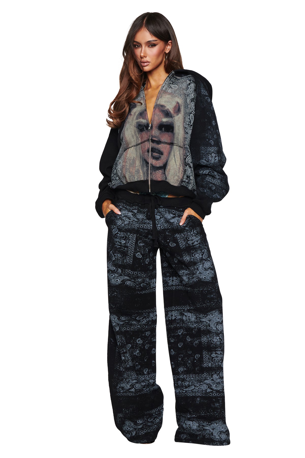 A woman stands with her hands in her pockets against a white background, exuding bold street style vibes in the I.AM.GIA T-REX ZIP THRU HOODIE - BLACK BANDANA and matching pants, featuring an oversized fit and a zip-up front adorned with a graphic of a blonde woman’s face.