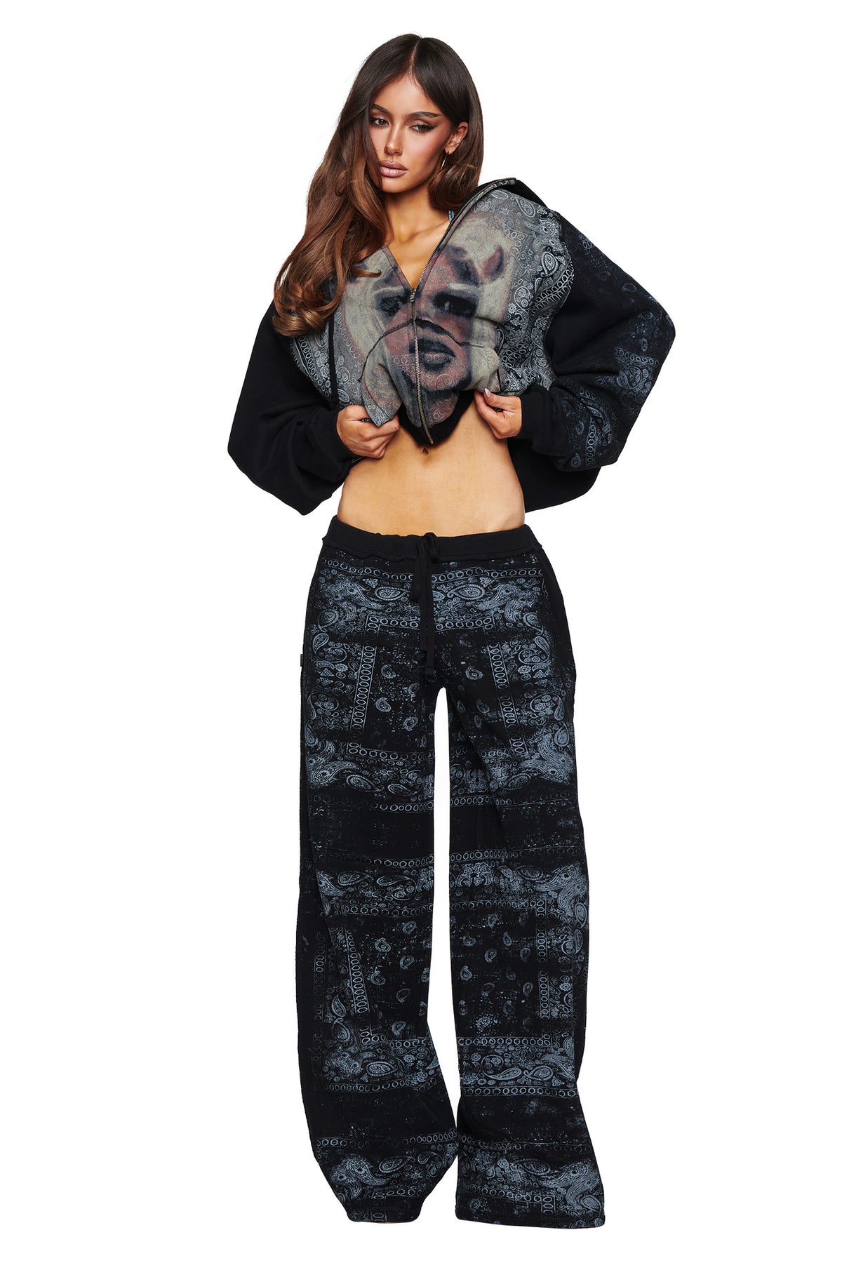 Against a white background, a woman showcases street style in the I.AM.GIA T-REX TRACKPANT - BLACK BANDANA, paired with a matching zip-up jacket featuring an eye-catching black bandana print and a graphic face on the front.