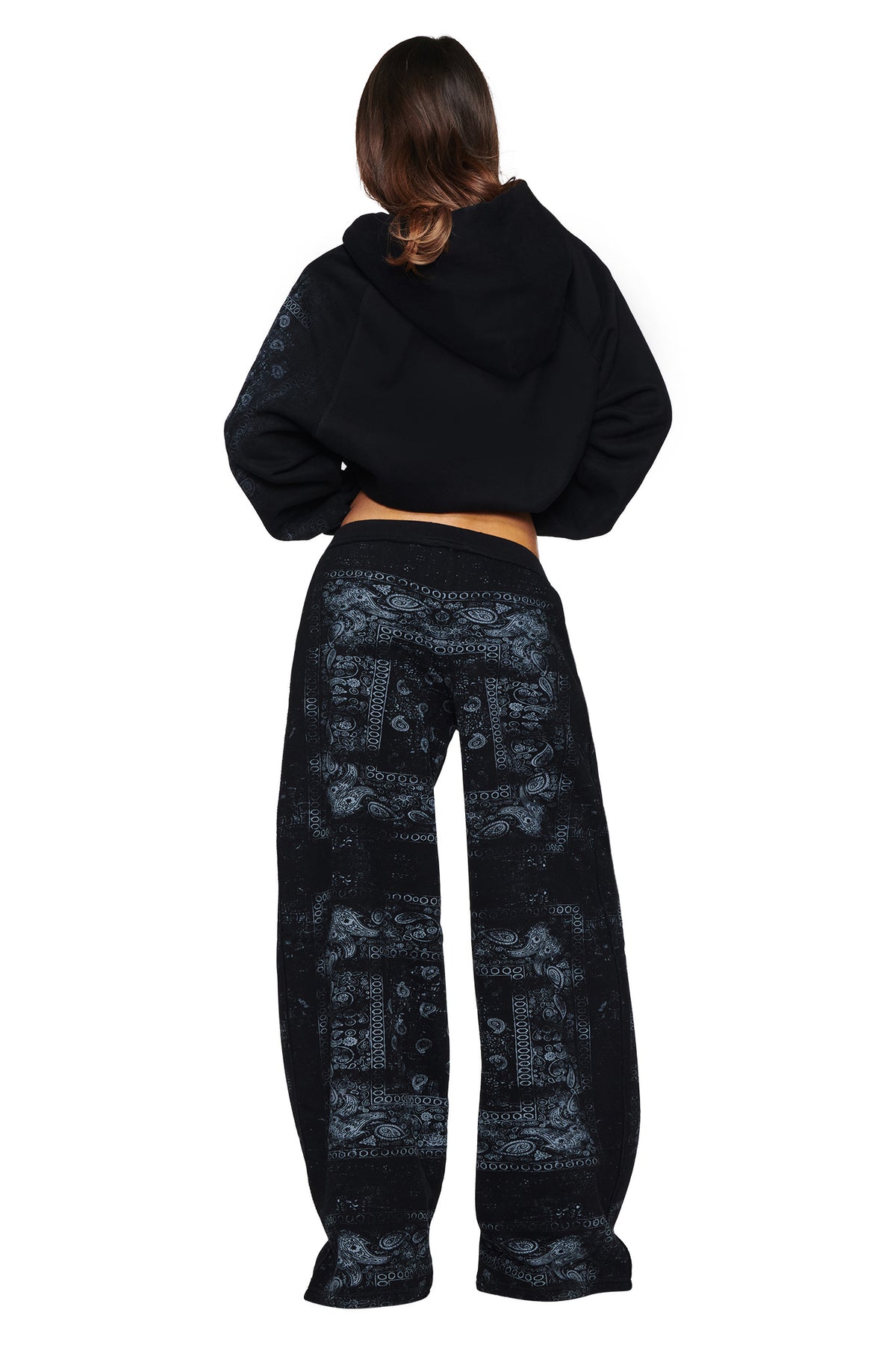 A person stands facing away, dressed in the I.AM.GIA T-REX ZIP THRU HOODIE - BLACK BANDANA and loose black pants with a paisley bandana pattern. The outfit embraces street style with its casual vibe, featuring the oversized zip-up hoodie against a clean, white background.
