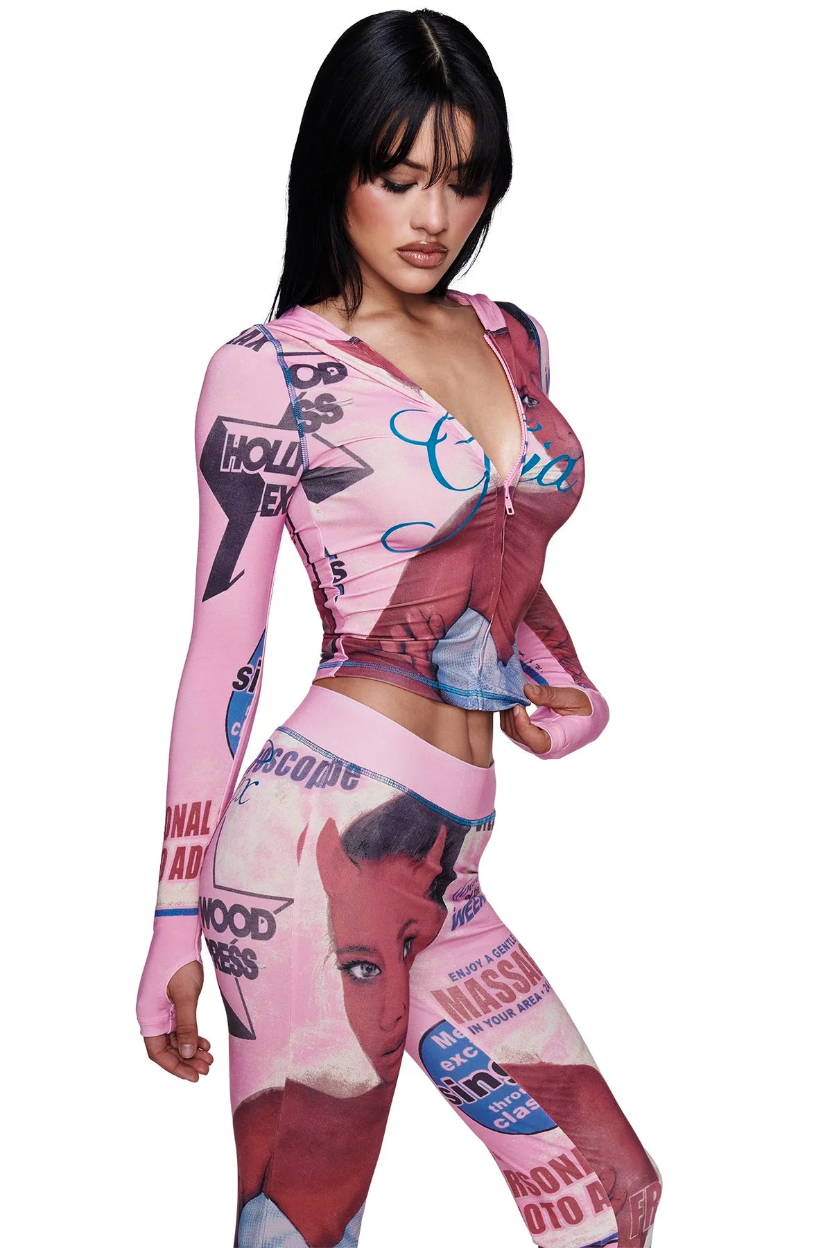 A woman wearing the I.AM.GIA T-REX BLARE TRACKPANT in pink and a coordinating pink top with retro comic-style graphics and "T-REX" text, looking down with her hands at her waist against a plain white background.