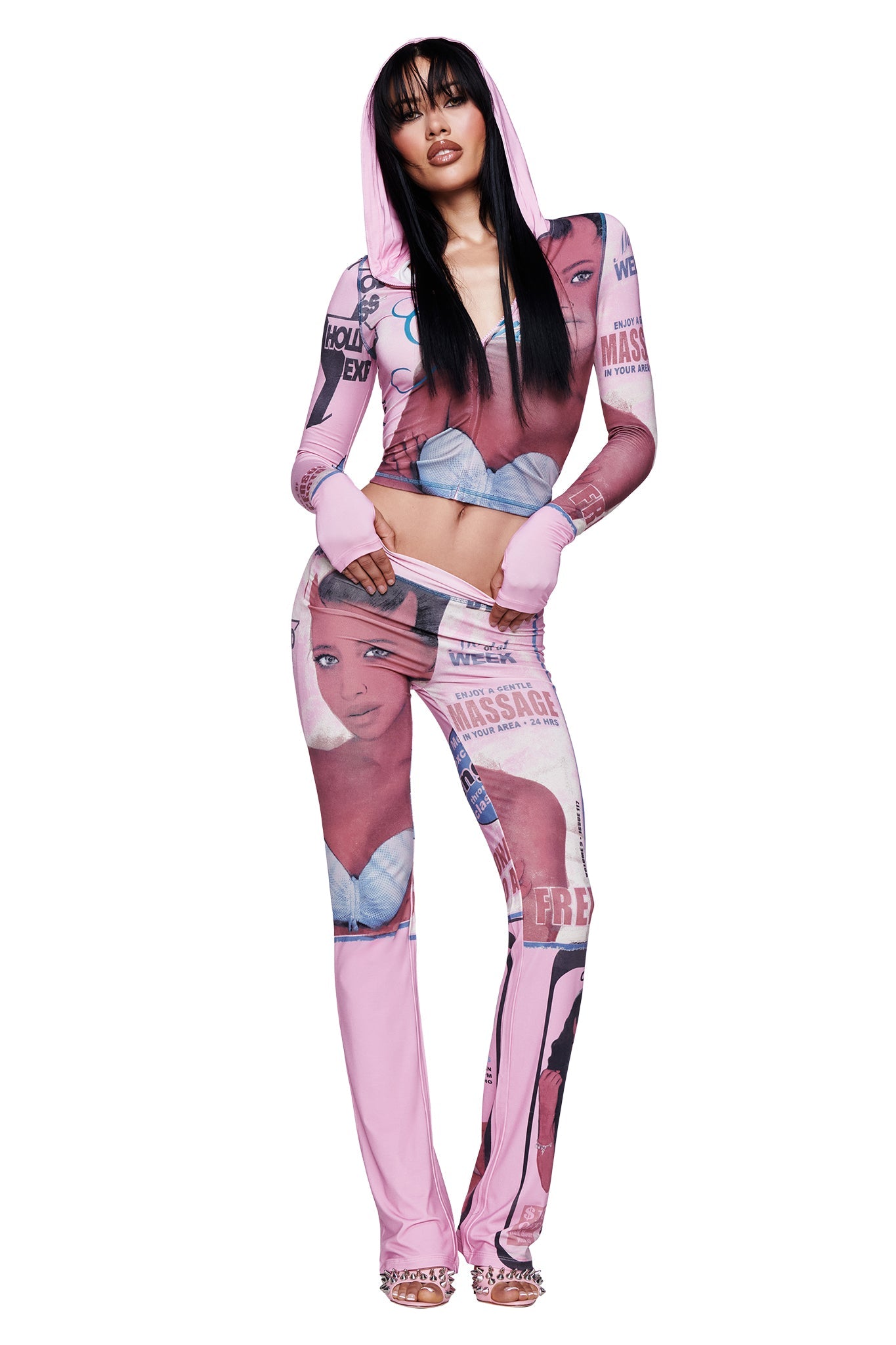 A woman poses in the T-REX BLARE HOODIE - PINK from I.AM.GIA with matching pants, both adorned with large printed graphics of a woman's face and bold lettering. She pairs the look with heels and wears her long dark hair in a style with bangs.