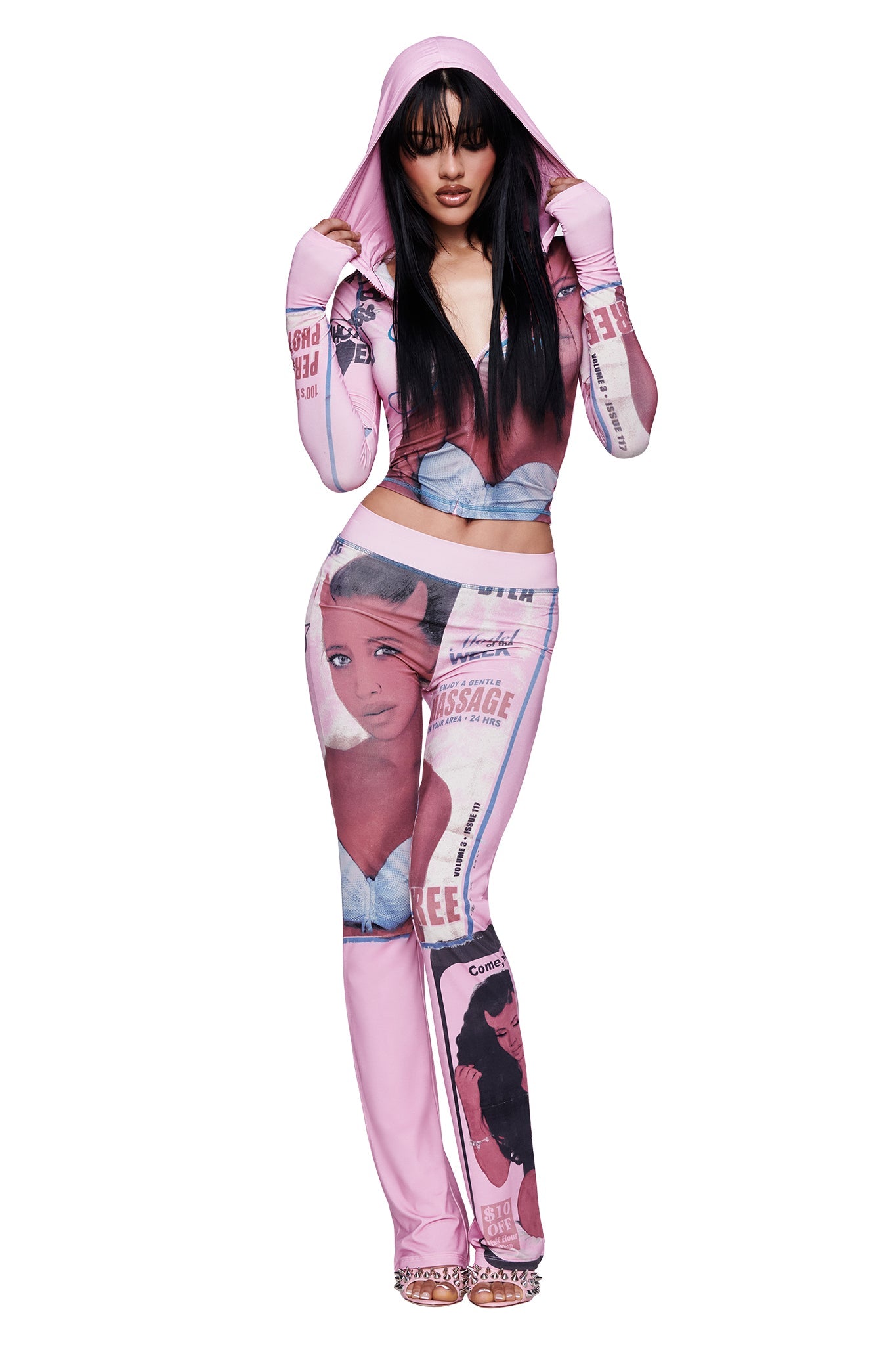 A woman poses with her hood up and hands on her head, showing off long dark hair with bangs, while wearing a pink hooded outfit featuring magazine cover and women’s images. She completes the look with the I.AM.GIA T-REX BLARE TRACKPANT in a stylish pink print.