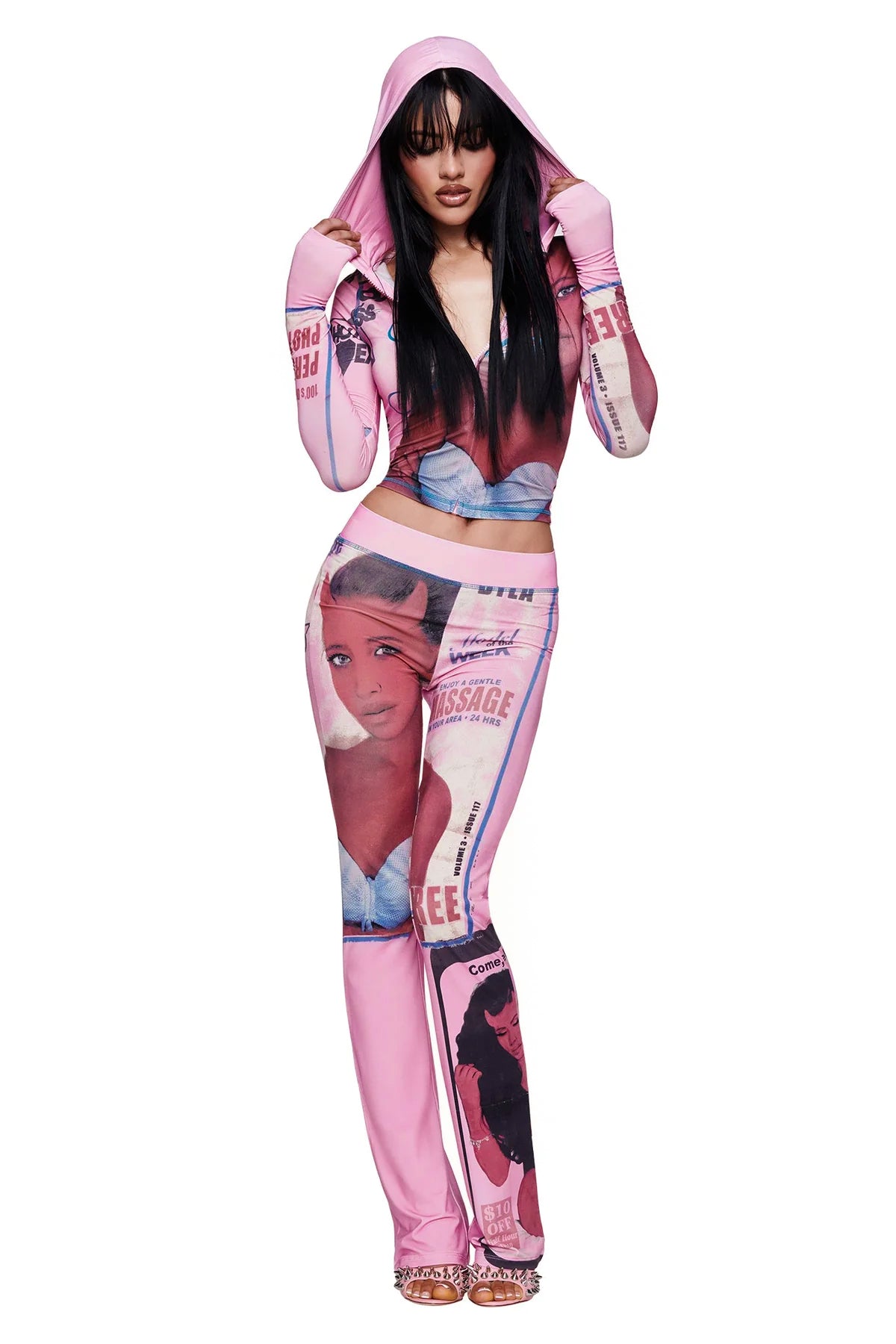 A woman wearing the I.AM.GIA T-REX BLARE TRACKPANT - PINK and a matching pink hooded crop top featuring T-REX pink print and graphic illustrated female faces poses with her hands on the hoodie, looking at the camera while standing against a white background.