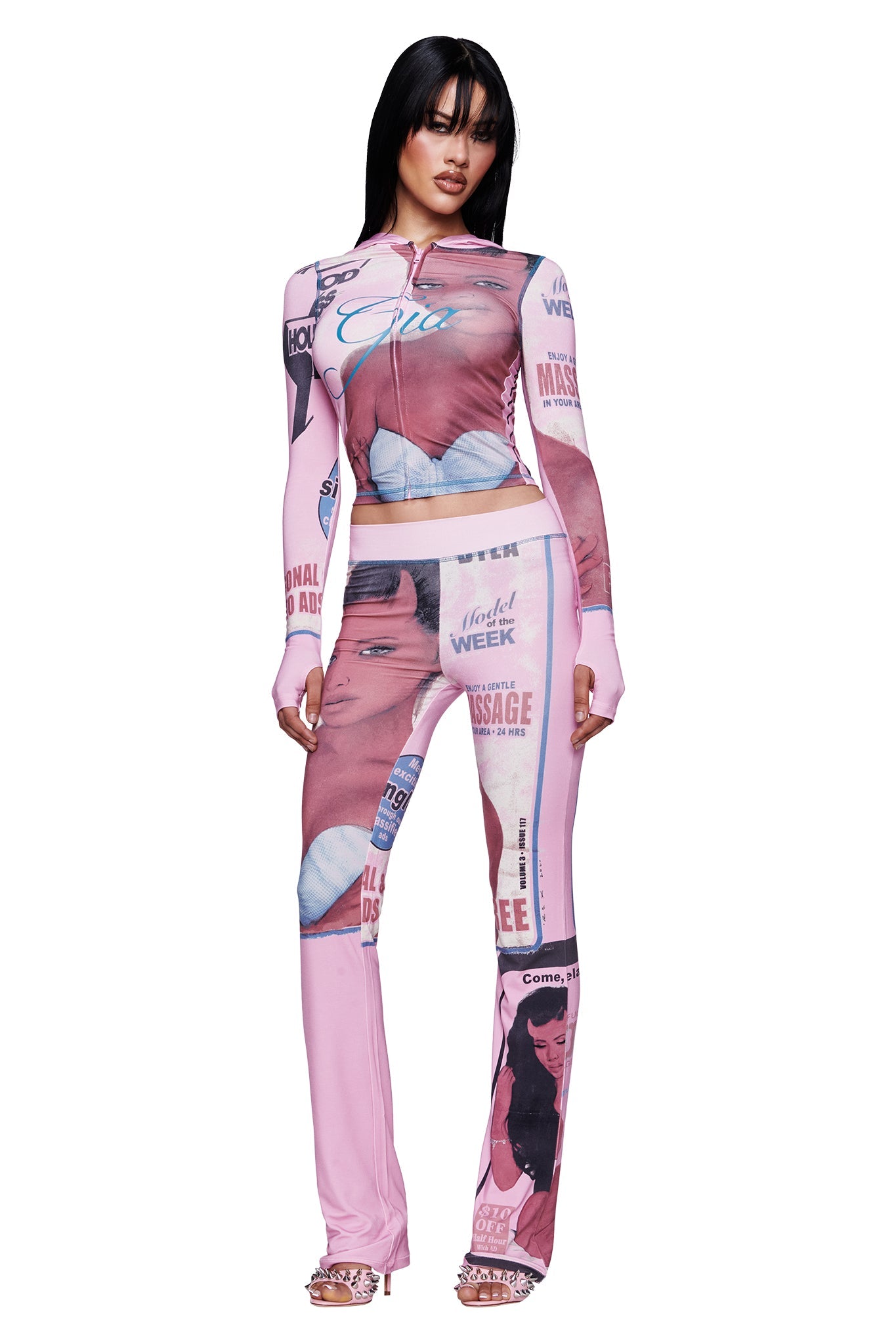 A woman stands in a pink, form-fitting outfit adorned with vintage magazine-style illustrations and text, featuring faces, lips, and various graphic elements. Her look is reminiscent of the playful aesthetic of the I.AM.GIA T-REX BLARE HOODIE in Pink. She completes her outfit with open-toed heels and wears her straight black hair with bangs.