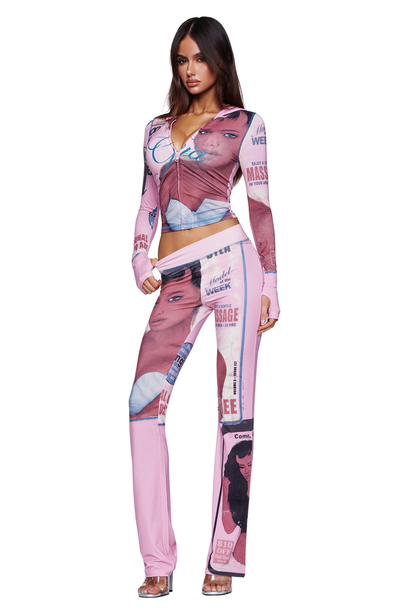 A woman stands wearing the I.AM.GIA T-REX BLARE HOODIE in pink, which features bold graphic prints of faces and magazine-inspired text. She pairs it with matching flared pants and poses confidently, one hand by her side and the other bent slightly, embodying an effortless blend of comfort and statement style.